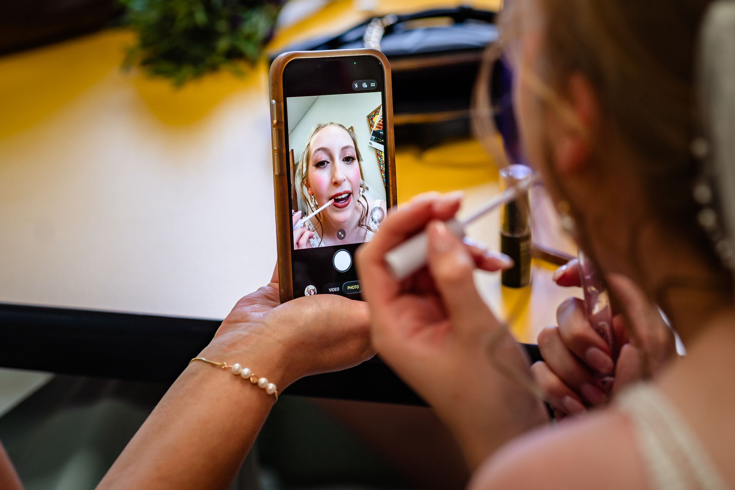 Bridesmaid photographs bride applying lipstick using a smartphone during wedding prep.