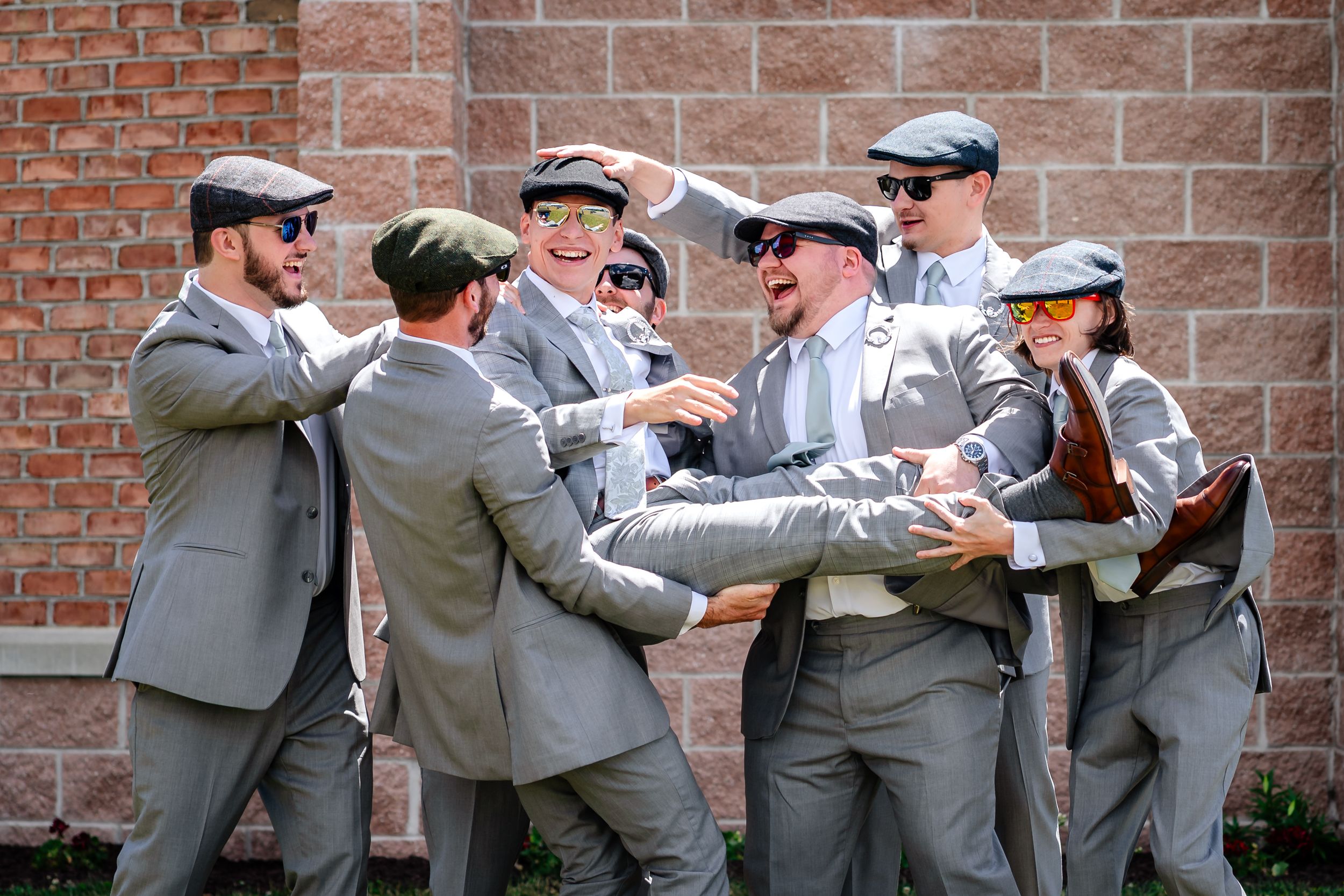 Groomsmen in gray suits and flat caps laughing and joking together outside brick building.