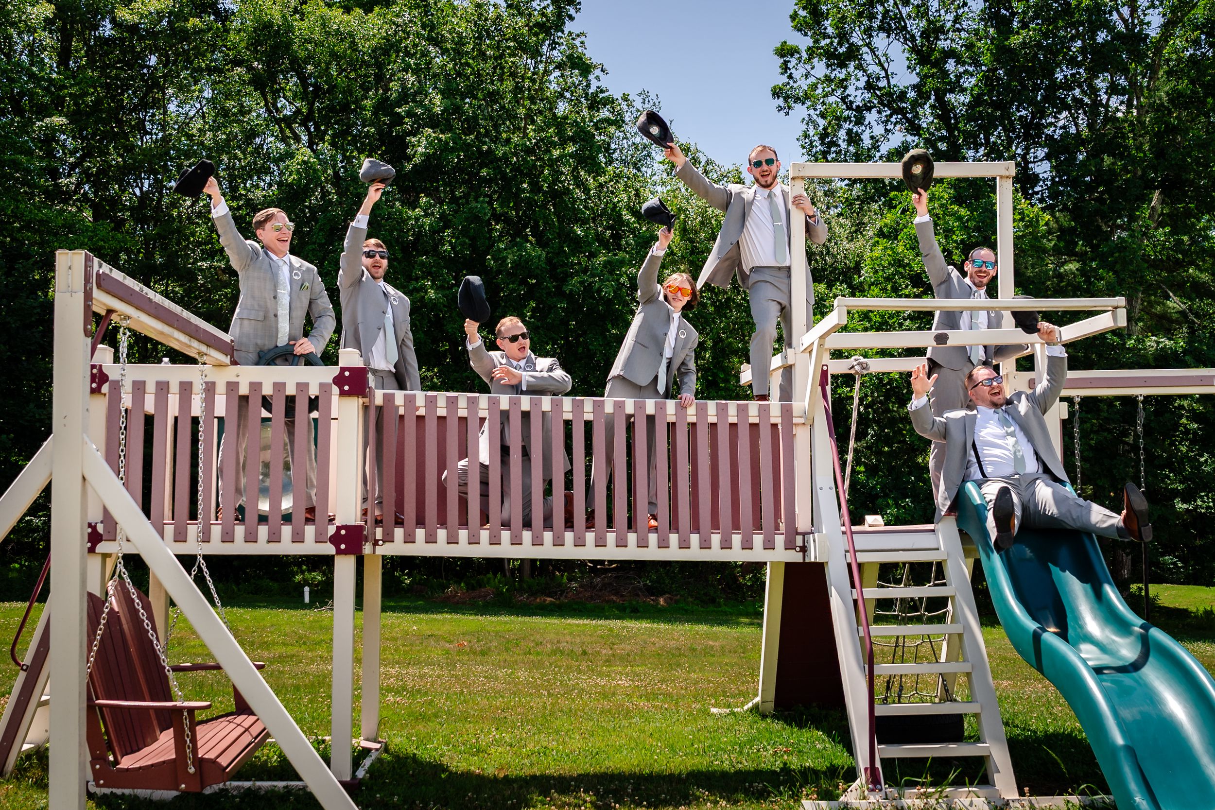 Groomsmen in gray suits celebrate on a backyard playground structure before a wedding.