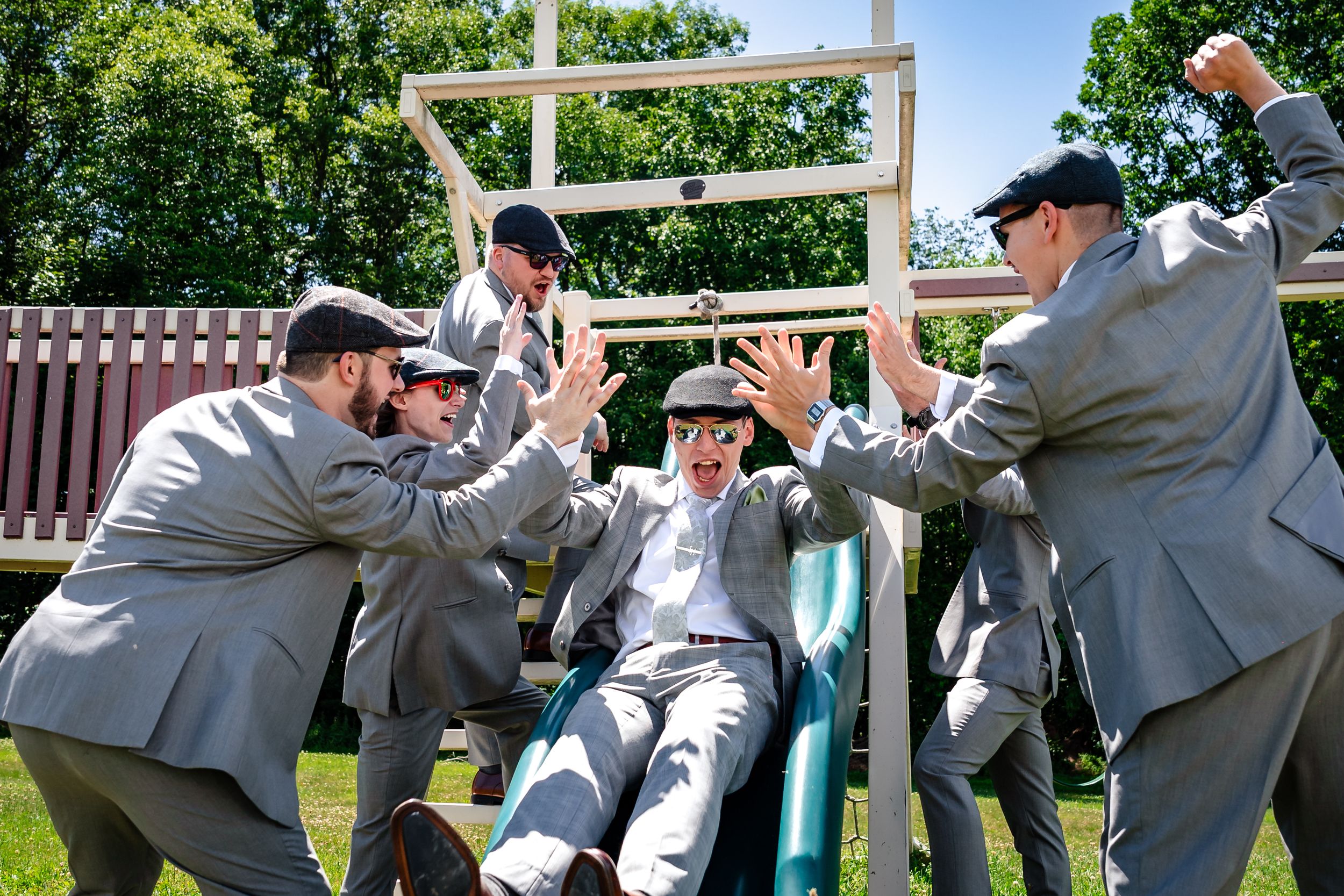 A group of men in matching gray suits celebrate on a playground slide on a sunny day.