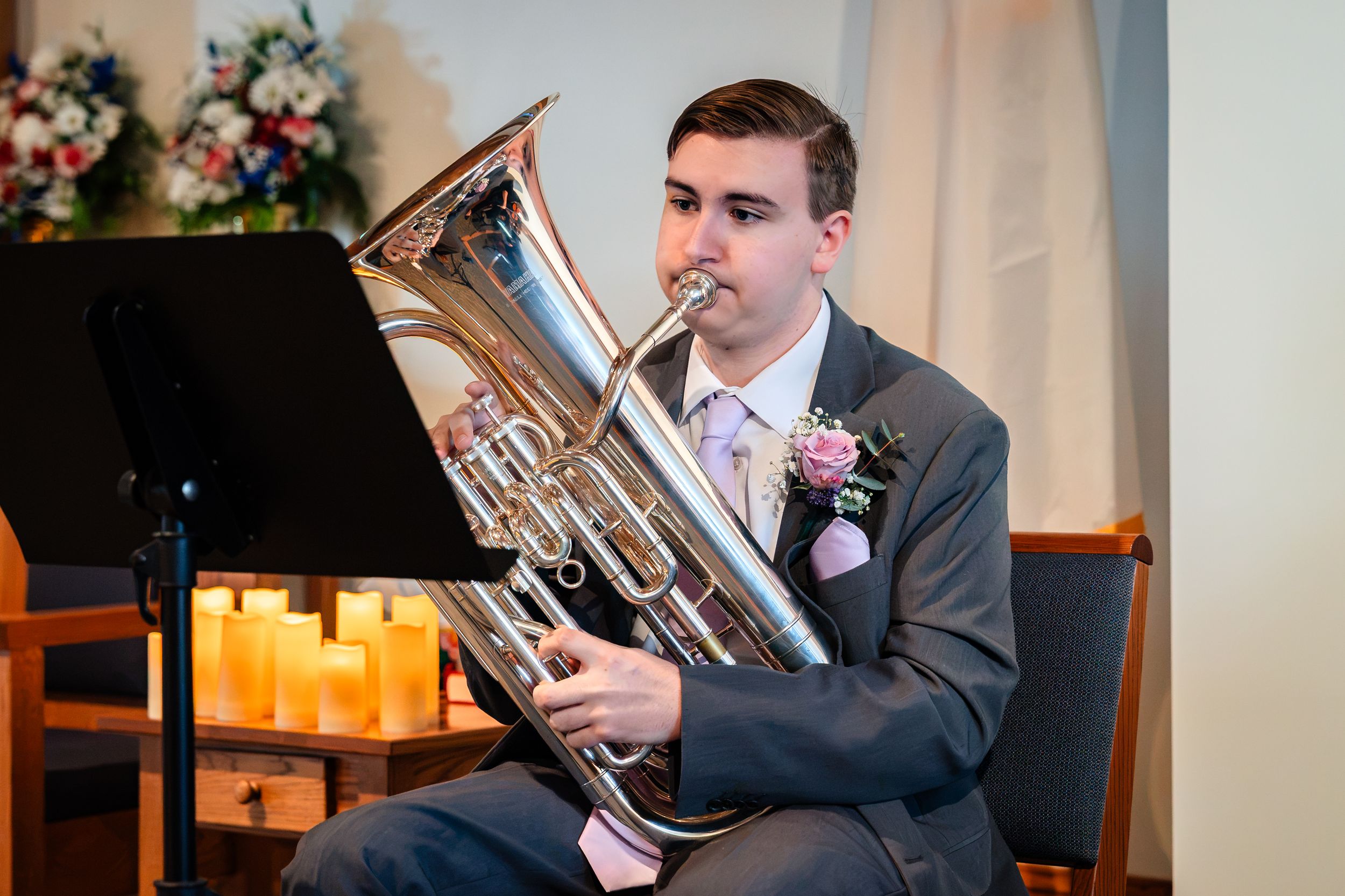 Groomsman plays euphonium during wedding ceremony beside candles and floral arrangements.