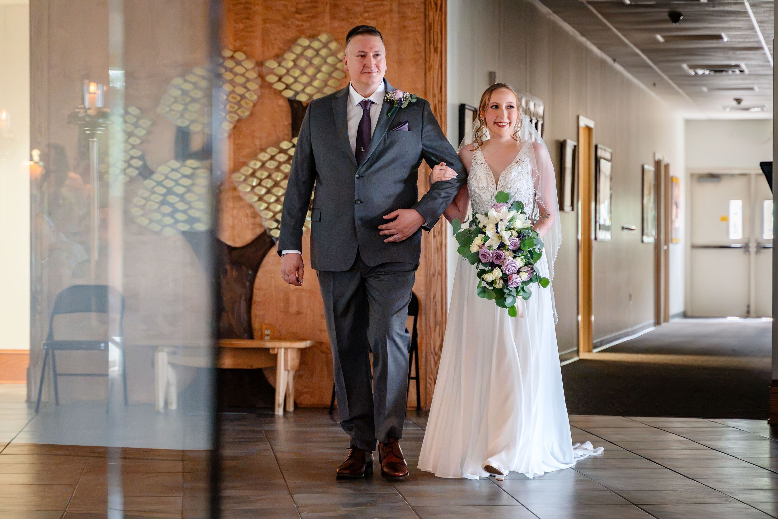 Bride holding floral bouquet walks with escort through venue lobby before ceremony.