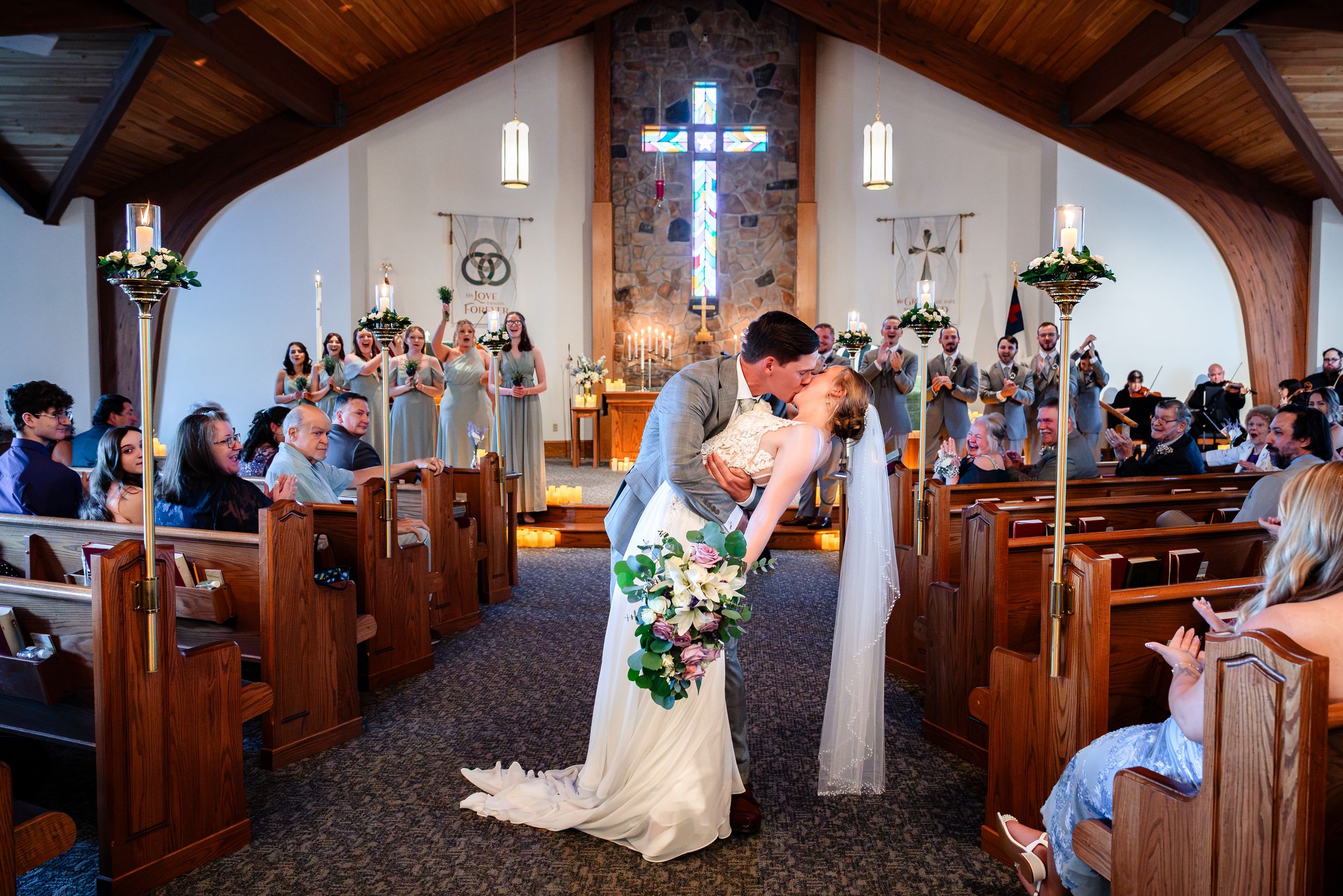 Newlyweds share romantic kiss in church aisle surrounded by cheering wedding guests.