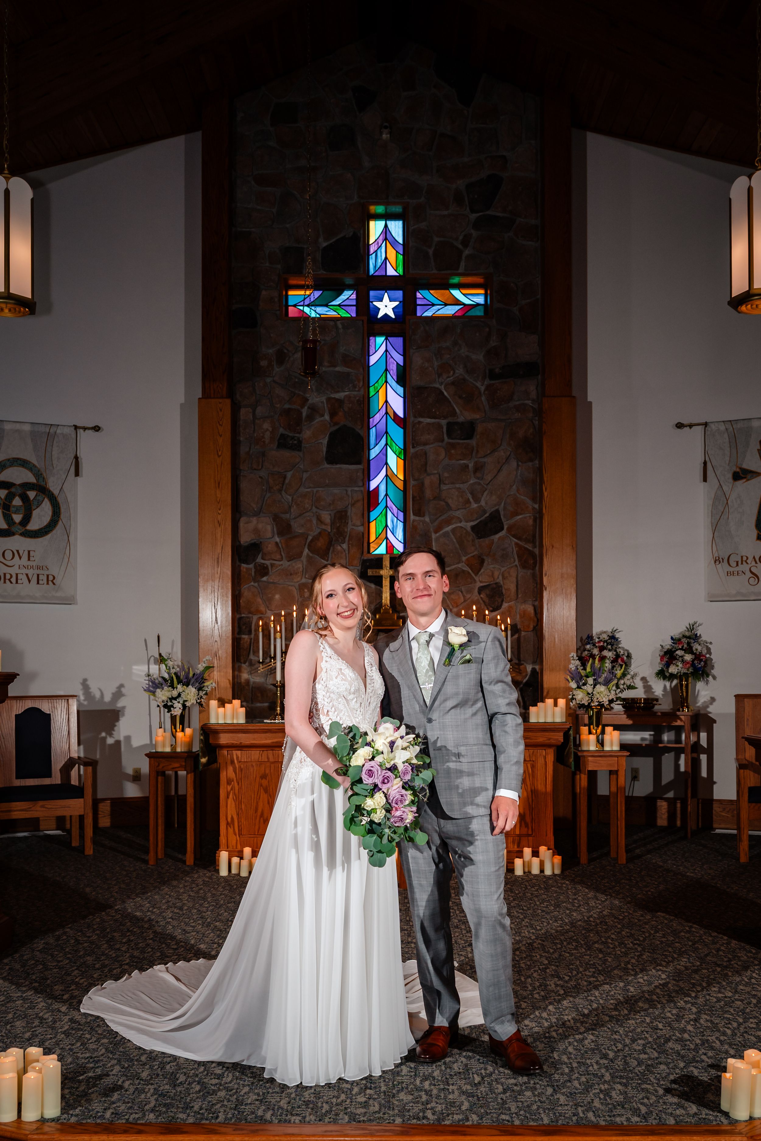 A bride and groom pose together at the altar of a church with a colorful stained glass cross.