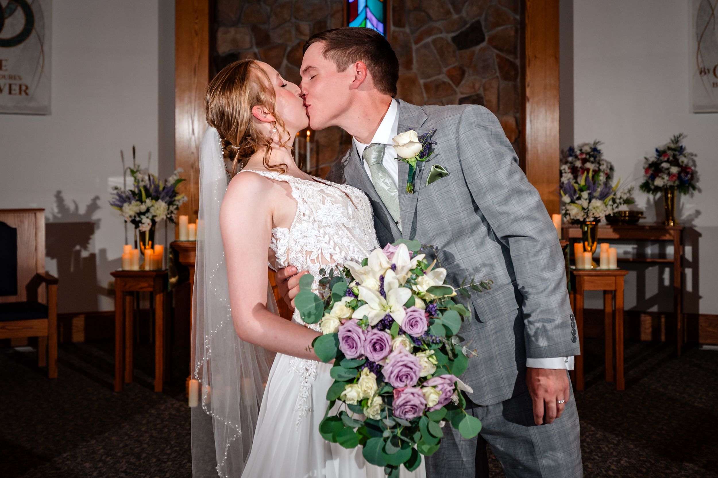 Bride and groom share first kiss at the altar in a candlelit stone chapel.