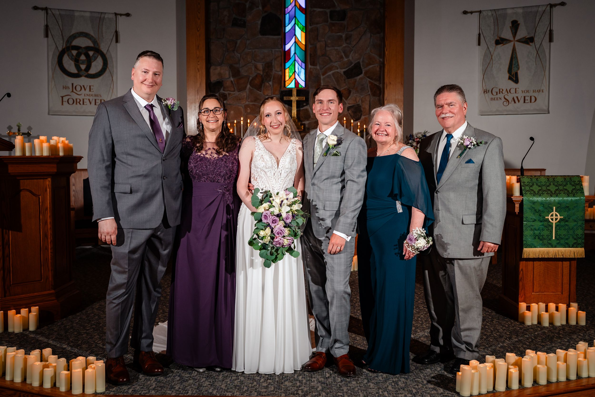 Bride and groom pose with four family members at church altar surrounded by candles after wedding ceremony.