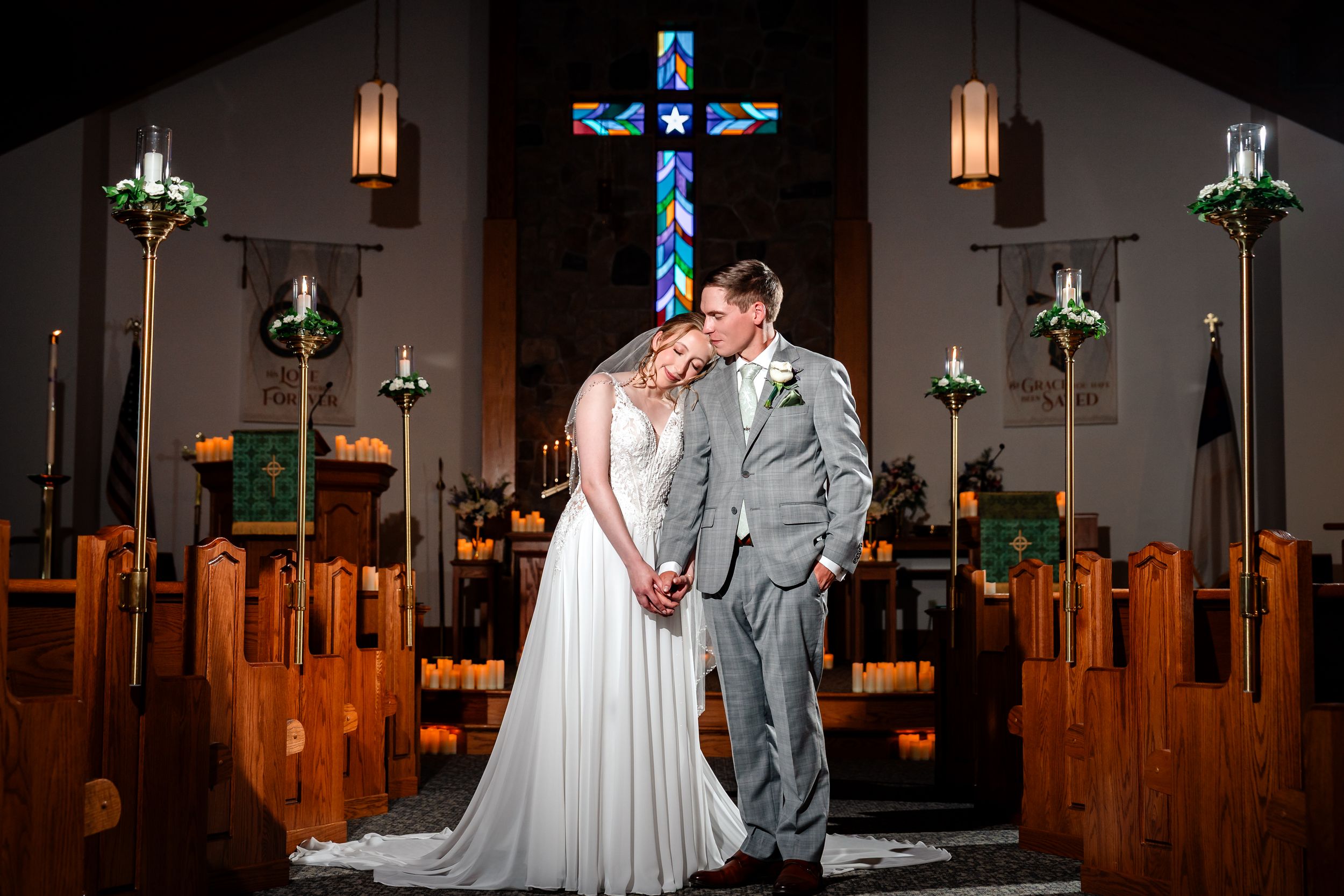 Bride and groom embrace before stunning stained glass cross at church altar.