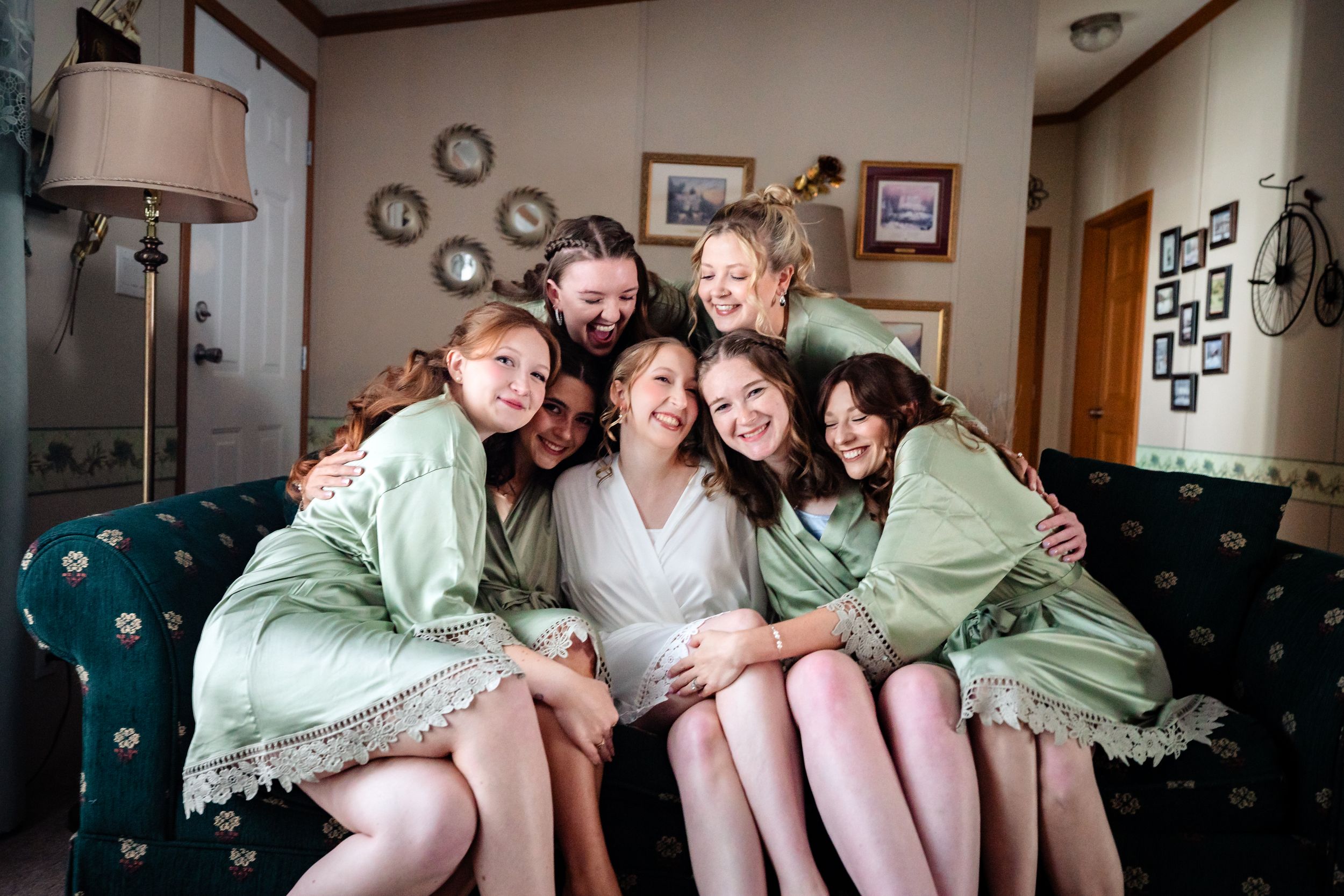 Bridesmaids in sage green robes huddle together laughing on a couch before the wedding.