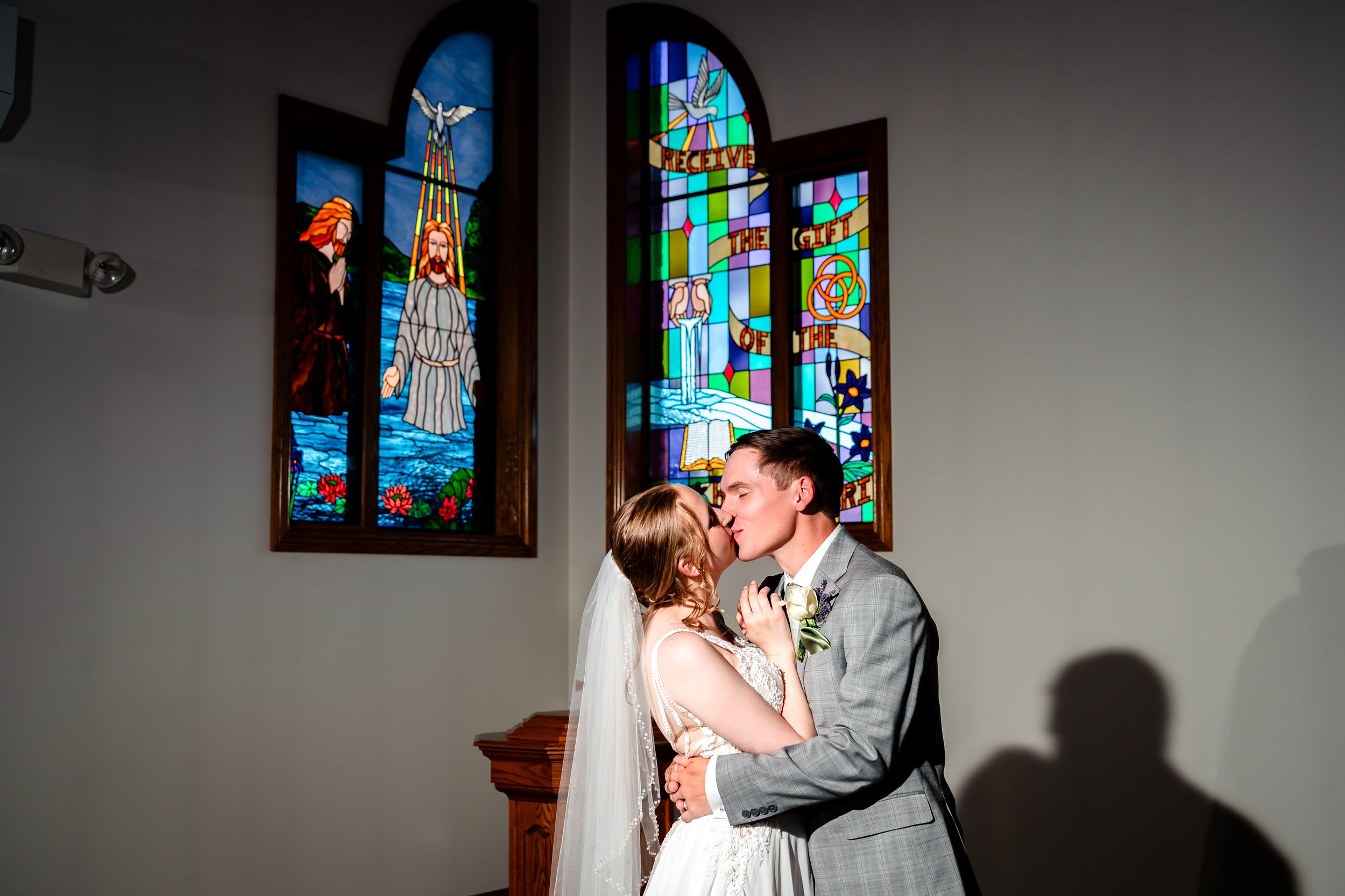 A newlywed couple shares their first kiss in front of stunning stained glass church windows.