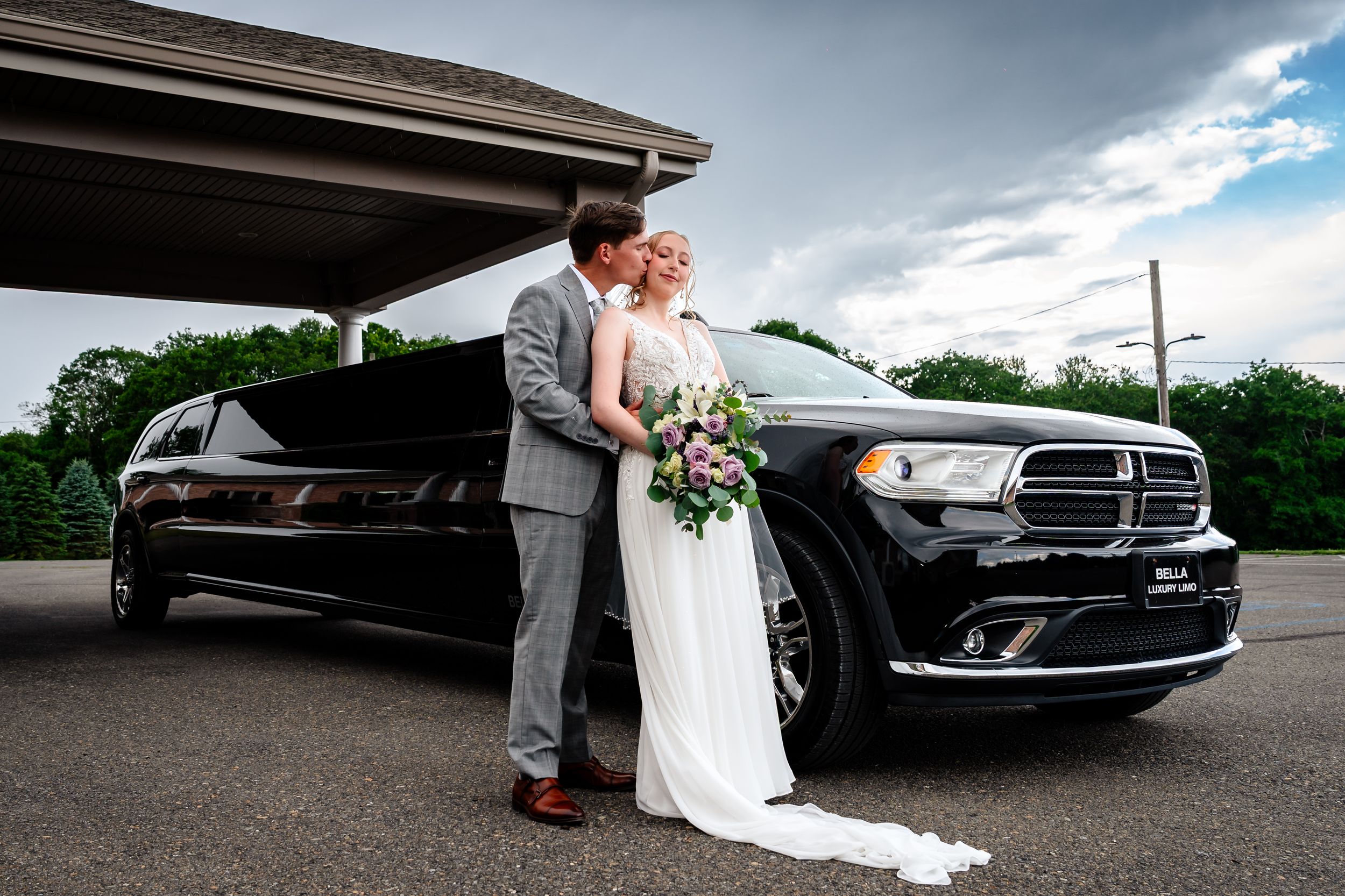 Bride and groom kissing in front of a black stretch Dodge Durango limousine on their wedding day.