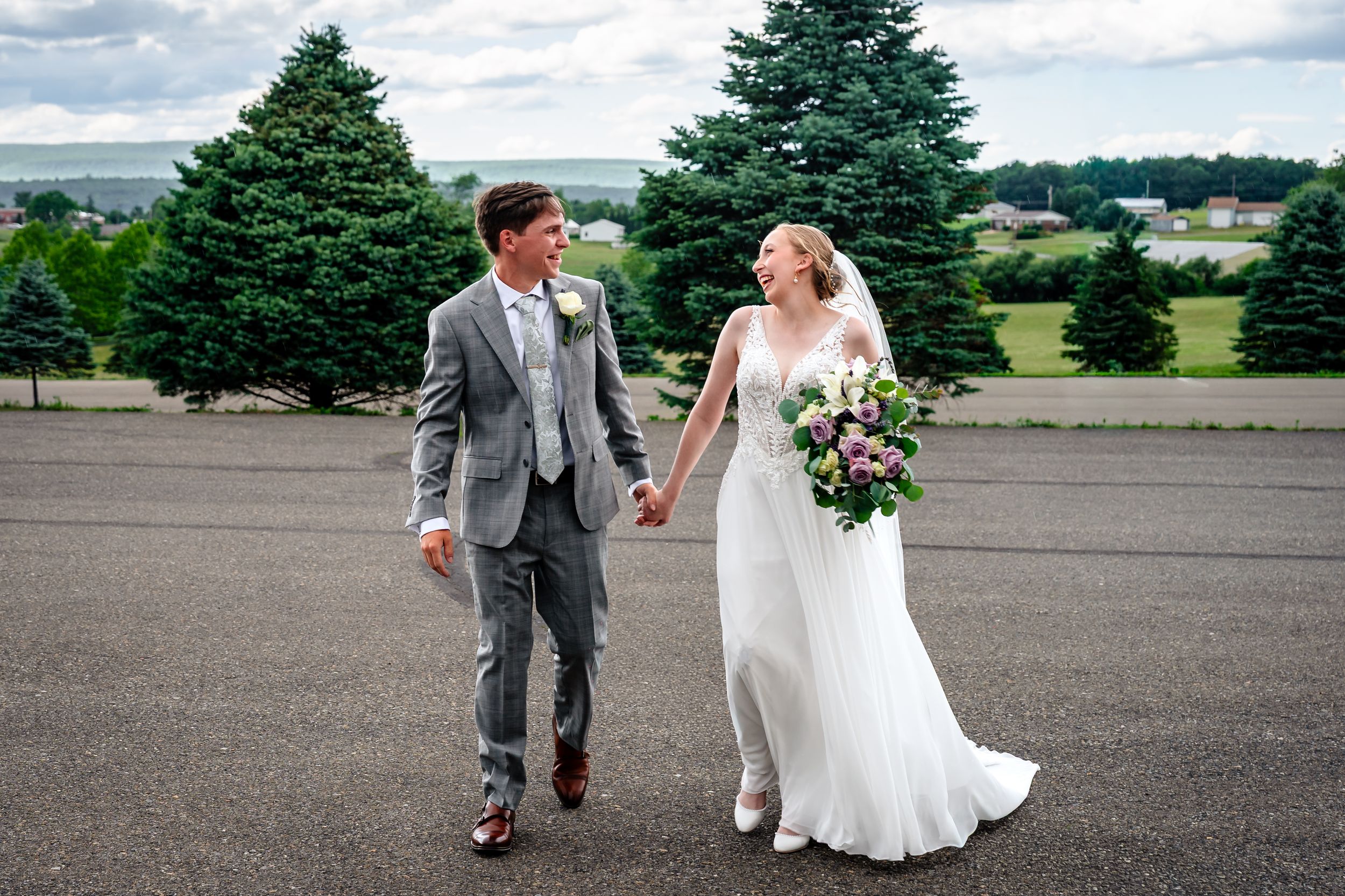 Bride and groom walk hand in hand on open tarmac with scenic countryside behind them.