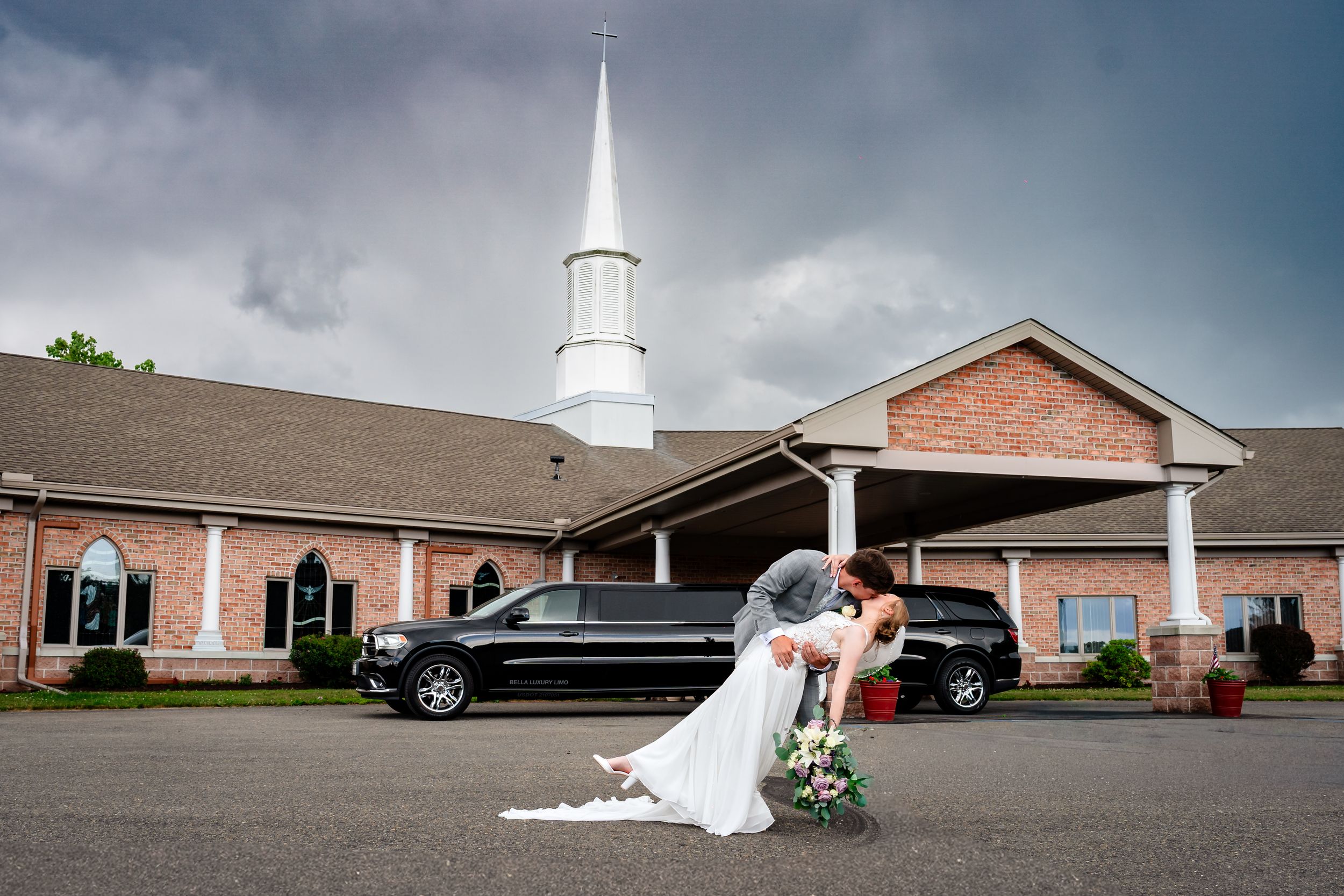 Groom dips bride dramatically outside brick church with limo and stormy sky behind.