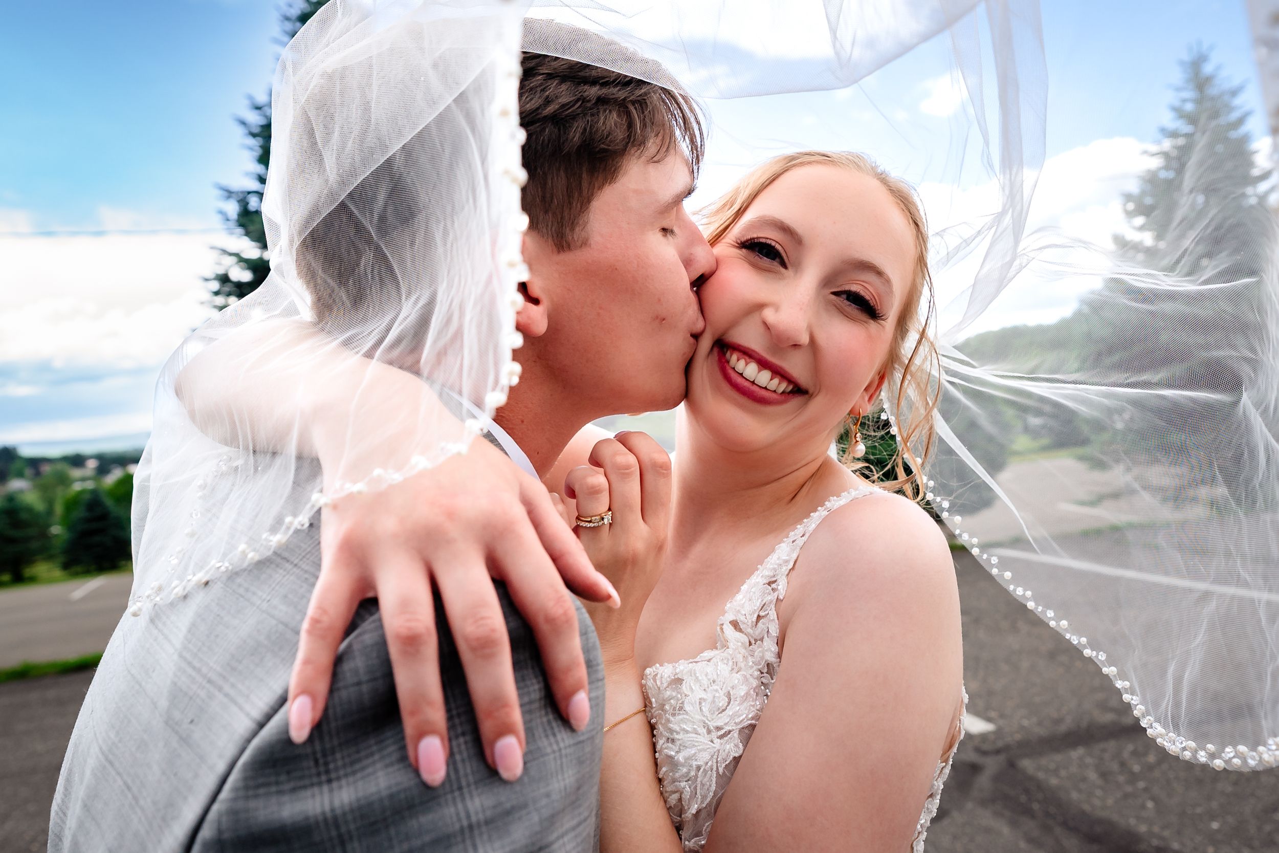 Groom kisses smiling bride on the cheek as her veil blows in the wind.