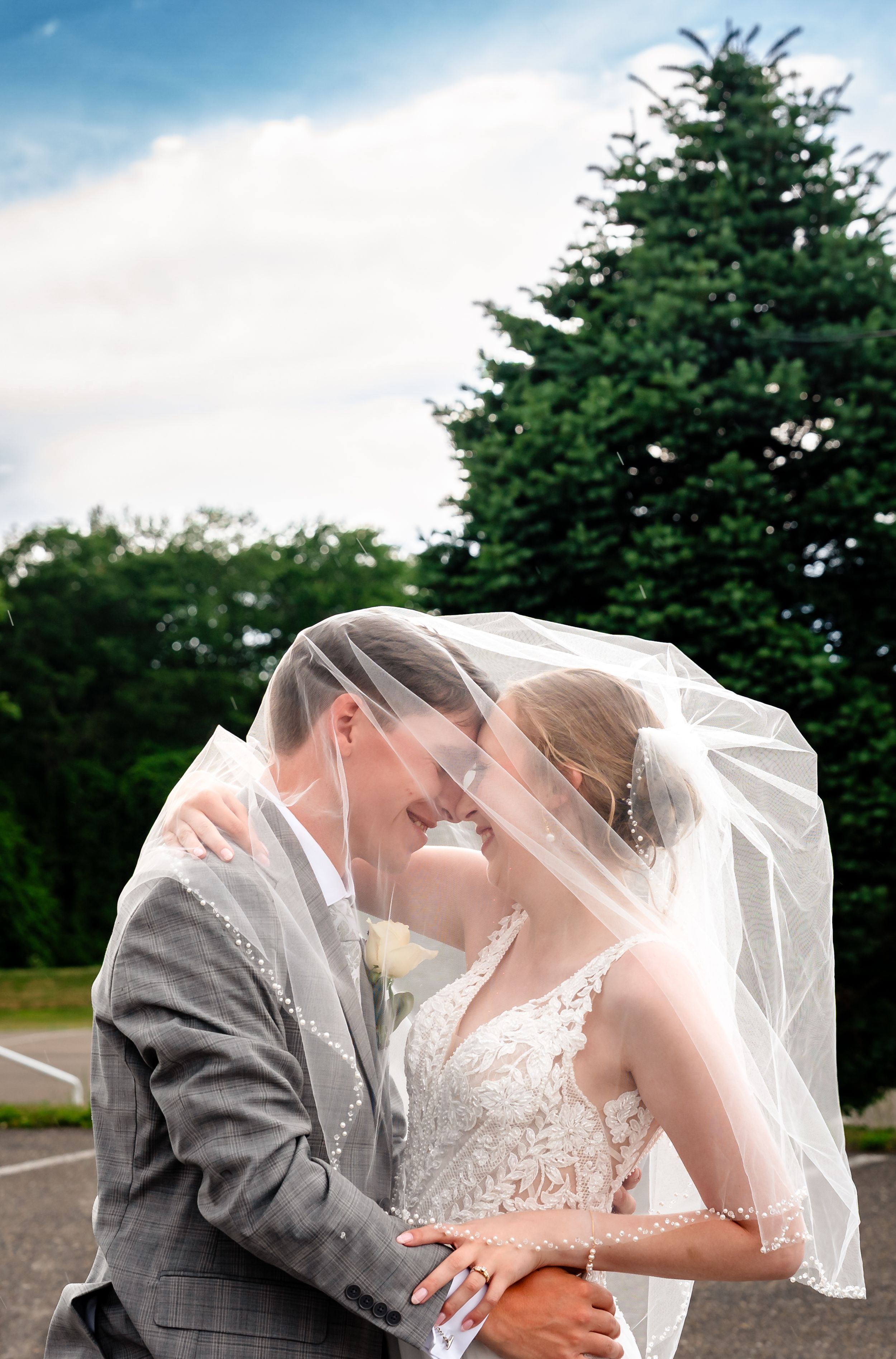 Bride and groom sharing a romantic moment under a wedding veil with lush green trees in the background.