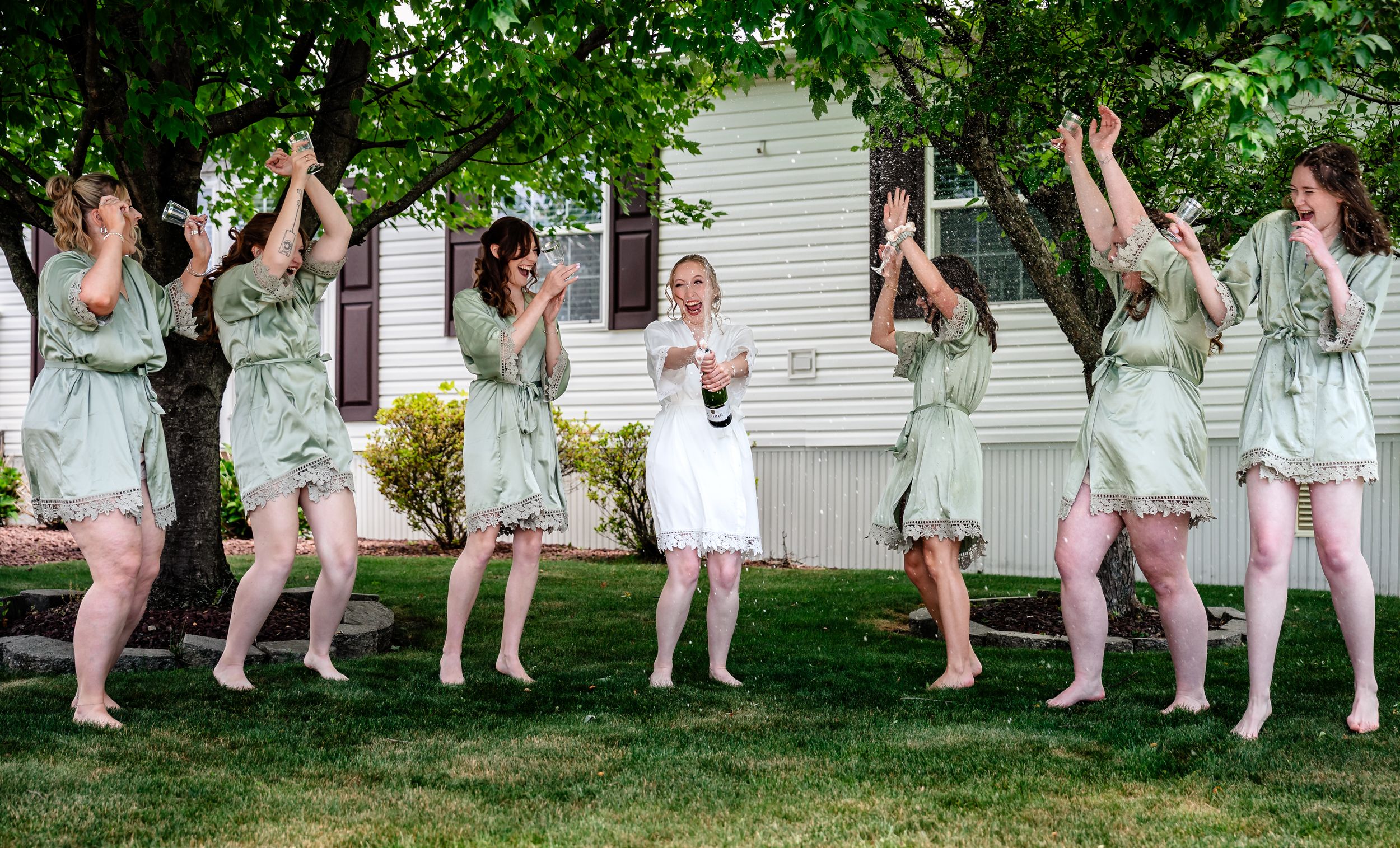 Bridesmaids in matching sage robes jump joyfully outdoors before a wedding ceremony.