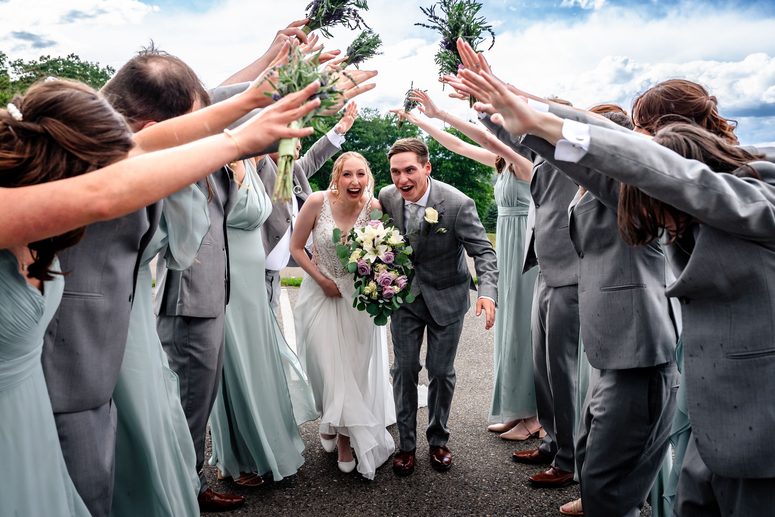 Bride and groom walk through an arch of raised hands formed by bridesmaids in sage green and groomsmen in gray suits.