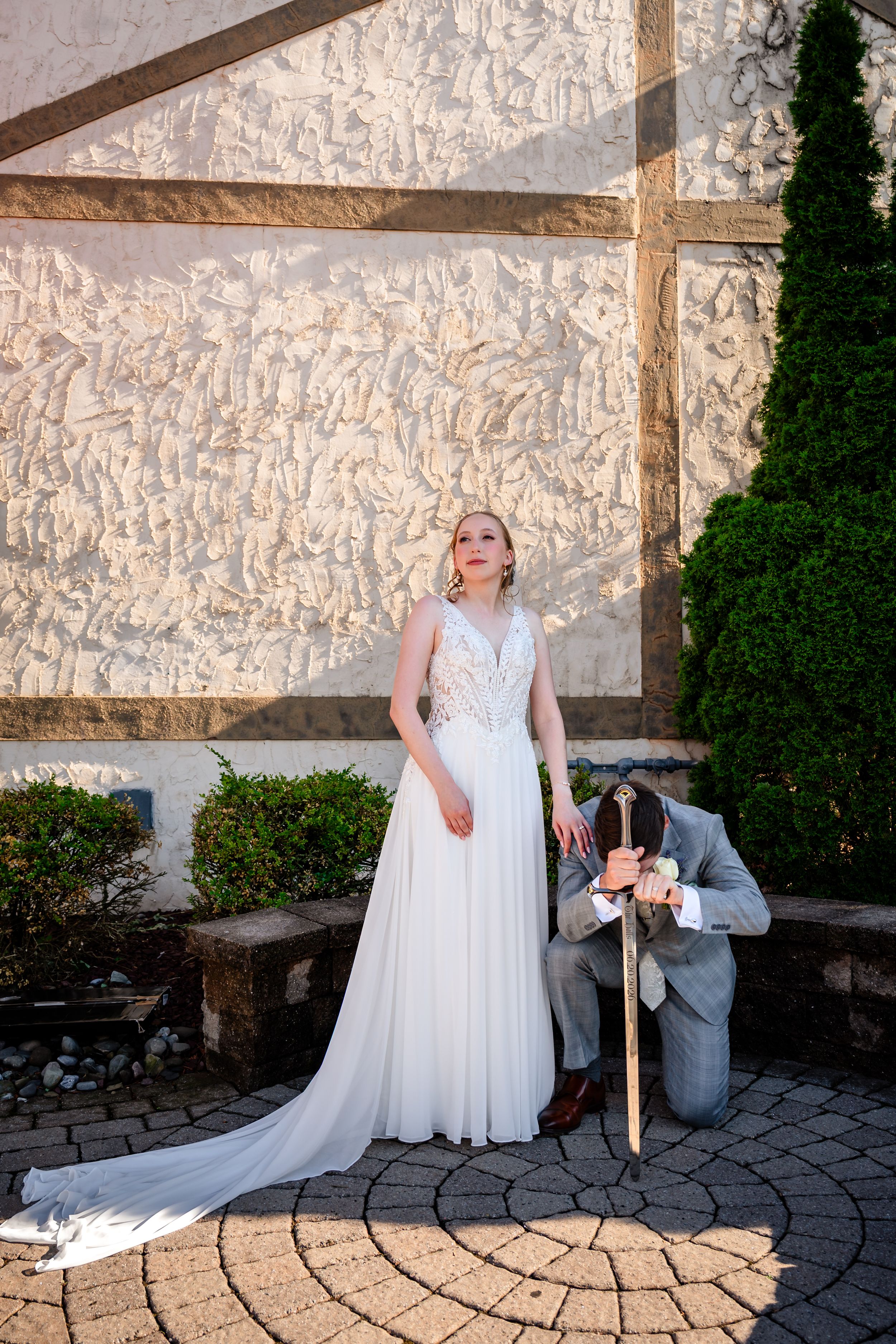 Bride in white gown stands elegantly while groom kneels beside her on a cobblestone patio.