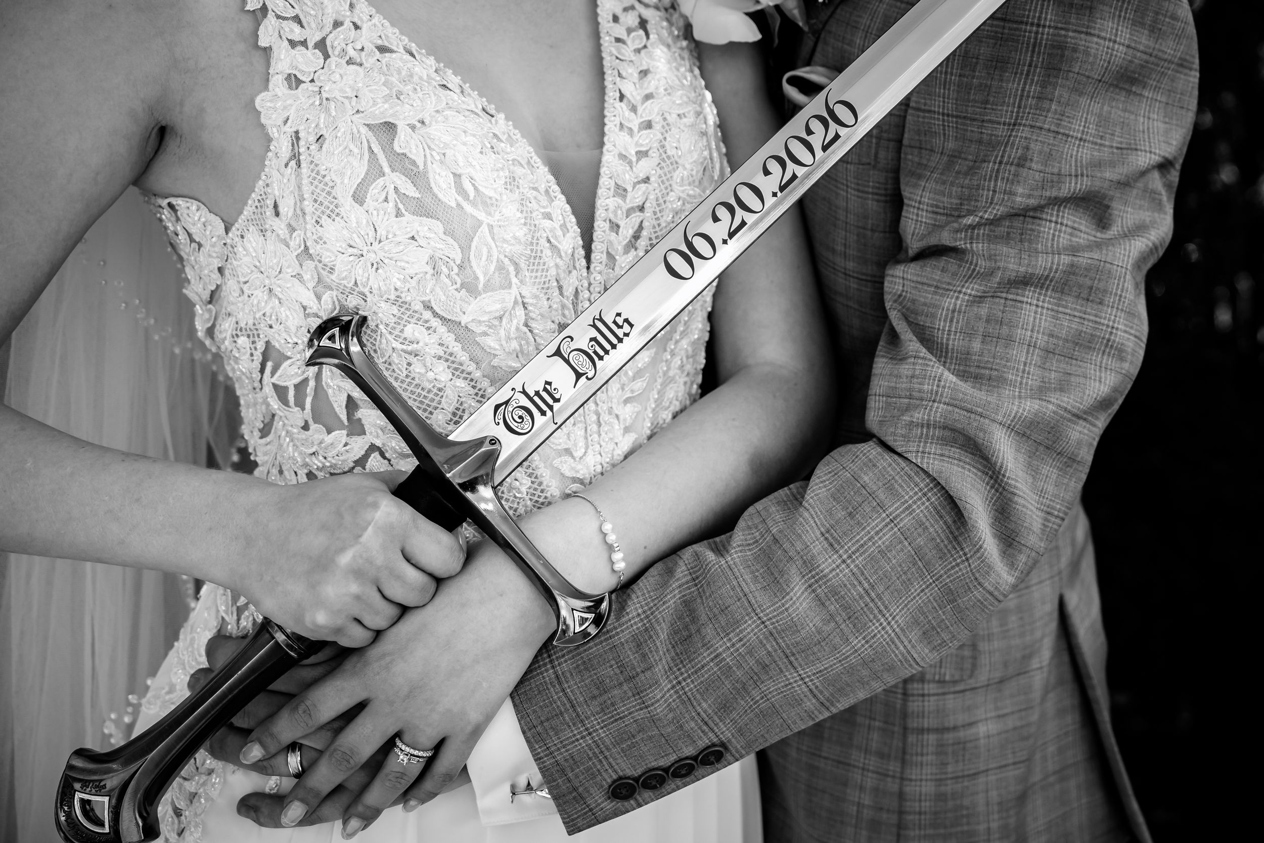 Black and white wedding photo of couple holding a sword engraved with The Hills and date 06.20.2020.