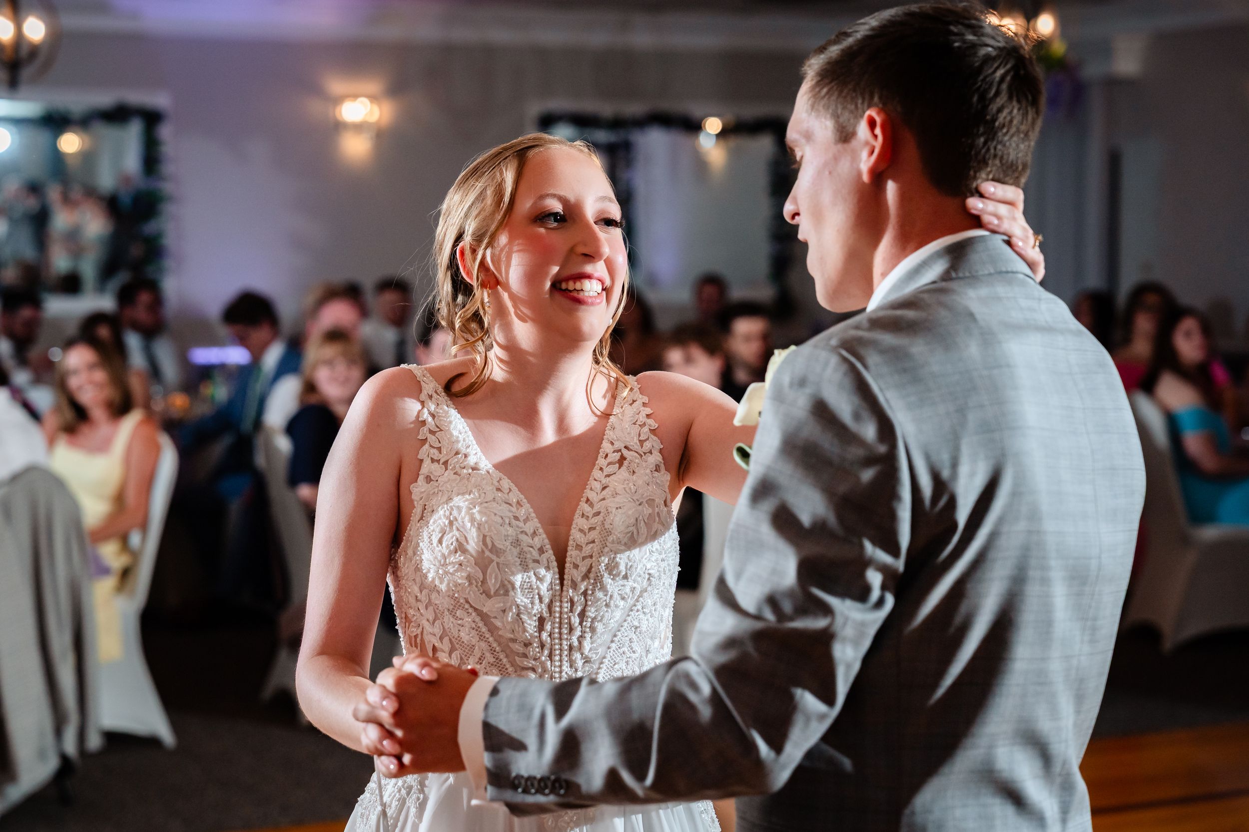 Bride and groom share their first dance at wedding reception, bride smiling in lace gown with guests seated in background.