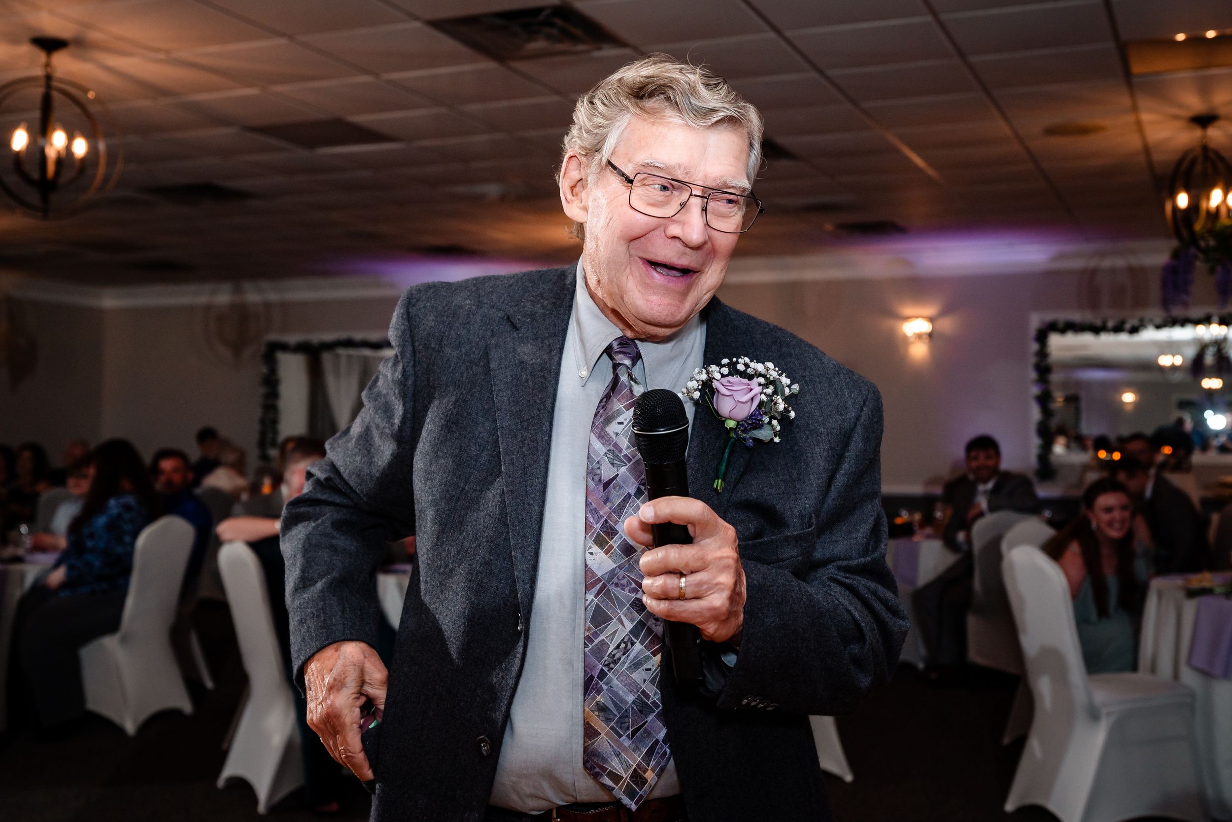 Elderly man smiling and speaking into a microphone at a wedding reception in a decorated banquet hall.