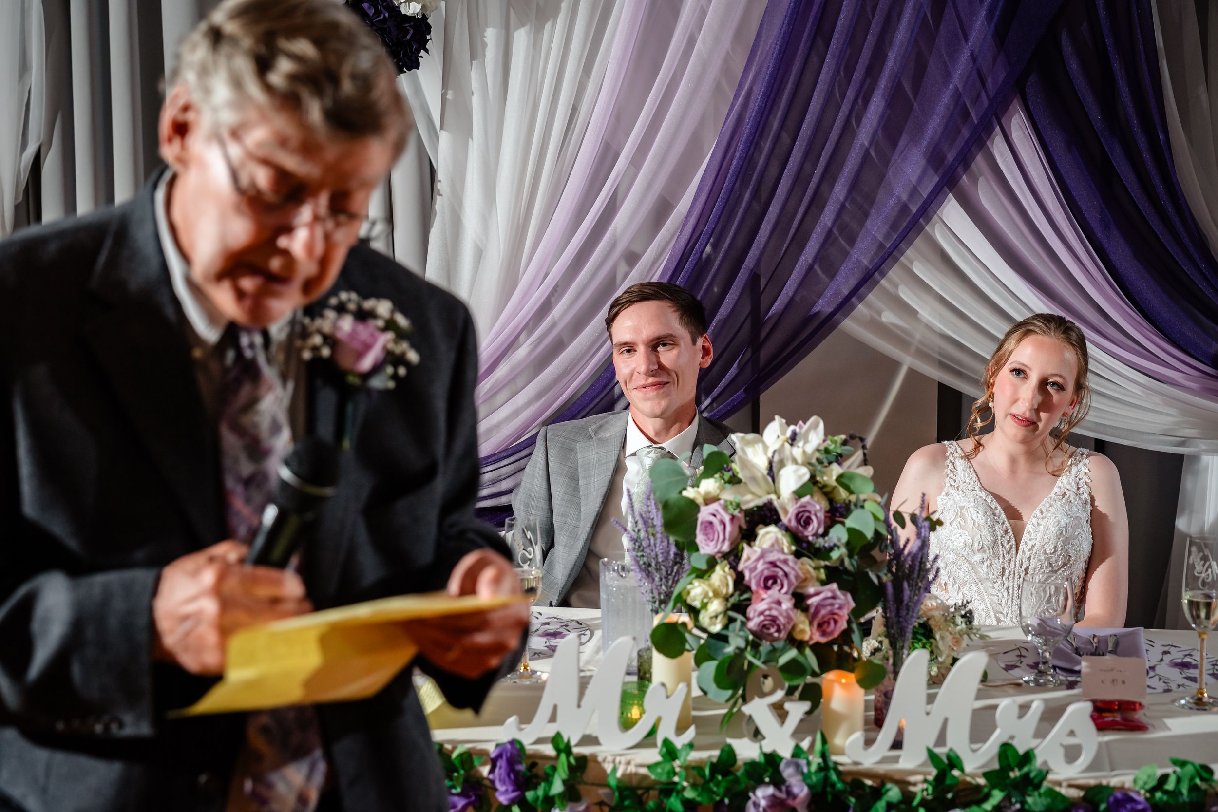 Elderly man reads speech from envelope at wedding reception as bride and groom listen at floral-decorated head table.