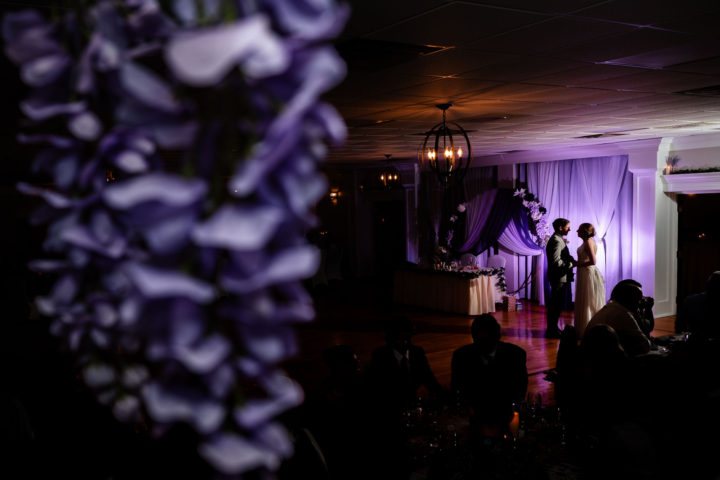 Couple shares first dance in elegant purple-lit wedding reception venue with floral decor.