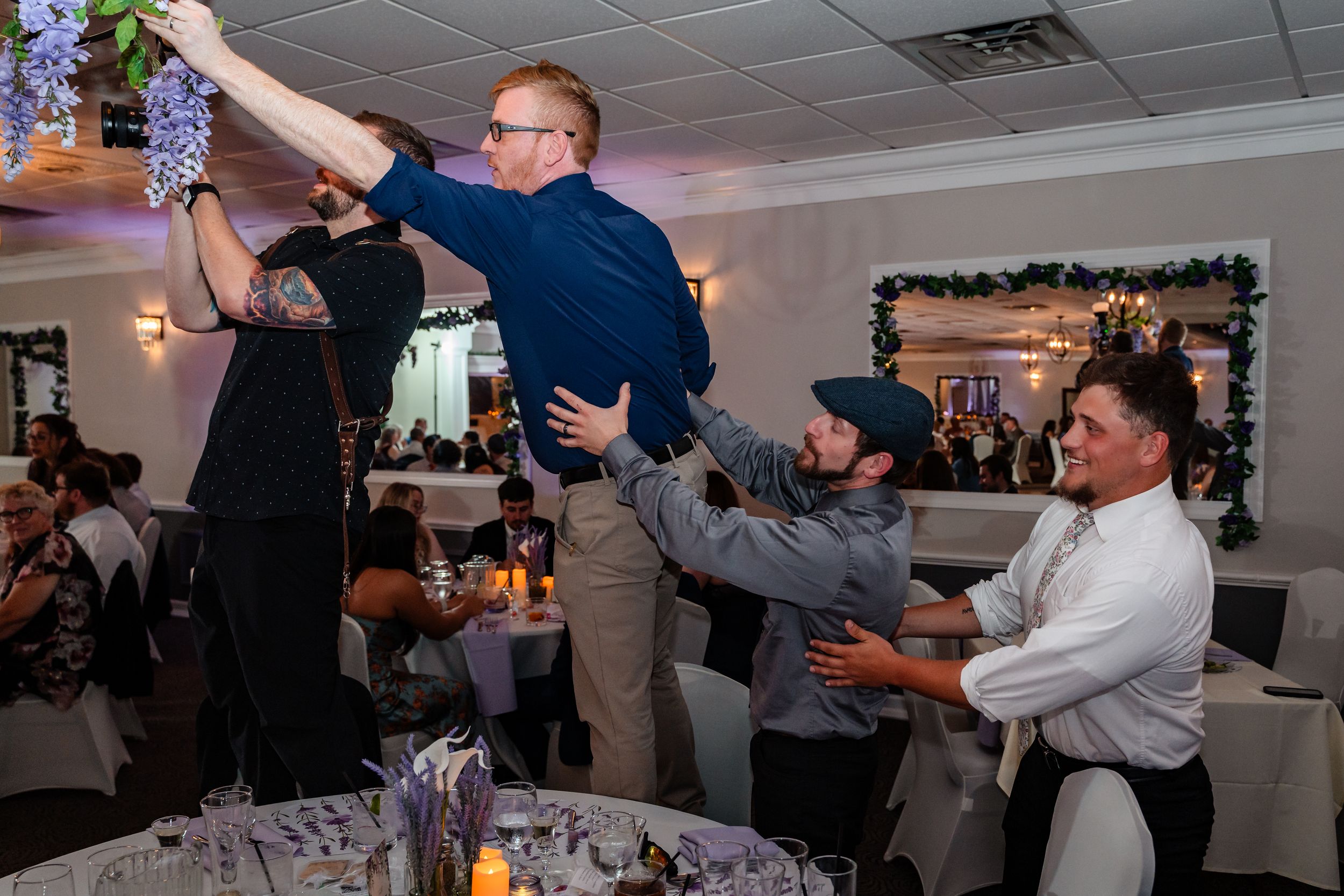 Groomsmen joyfully lift a guest during a lively wedding reception celebration.