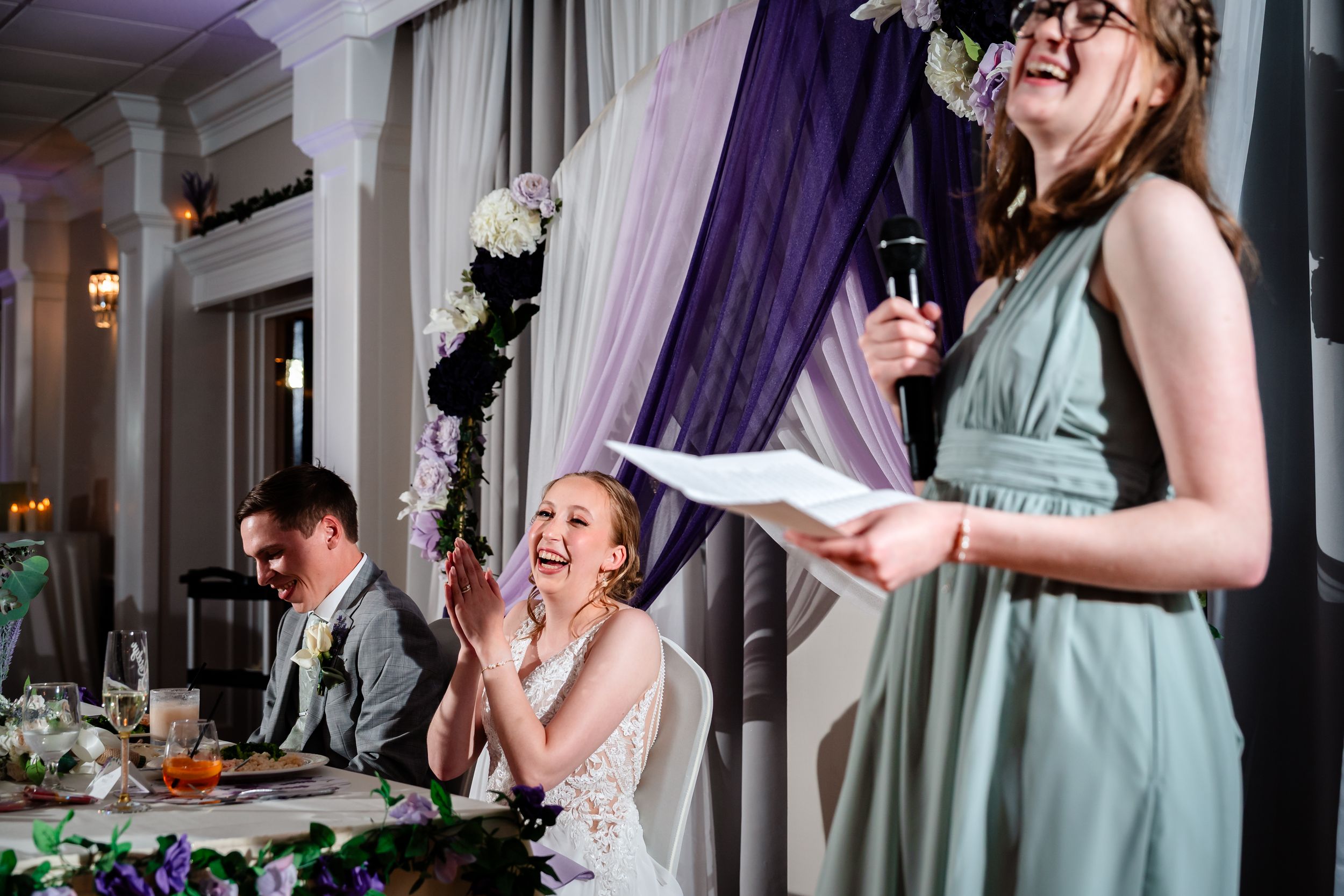 Bride laughs joyfully during wedding reception toast as bridesmaid reads speech at elegantly decorated venue.