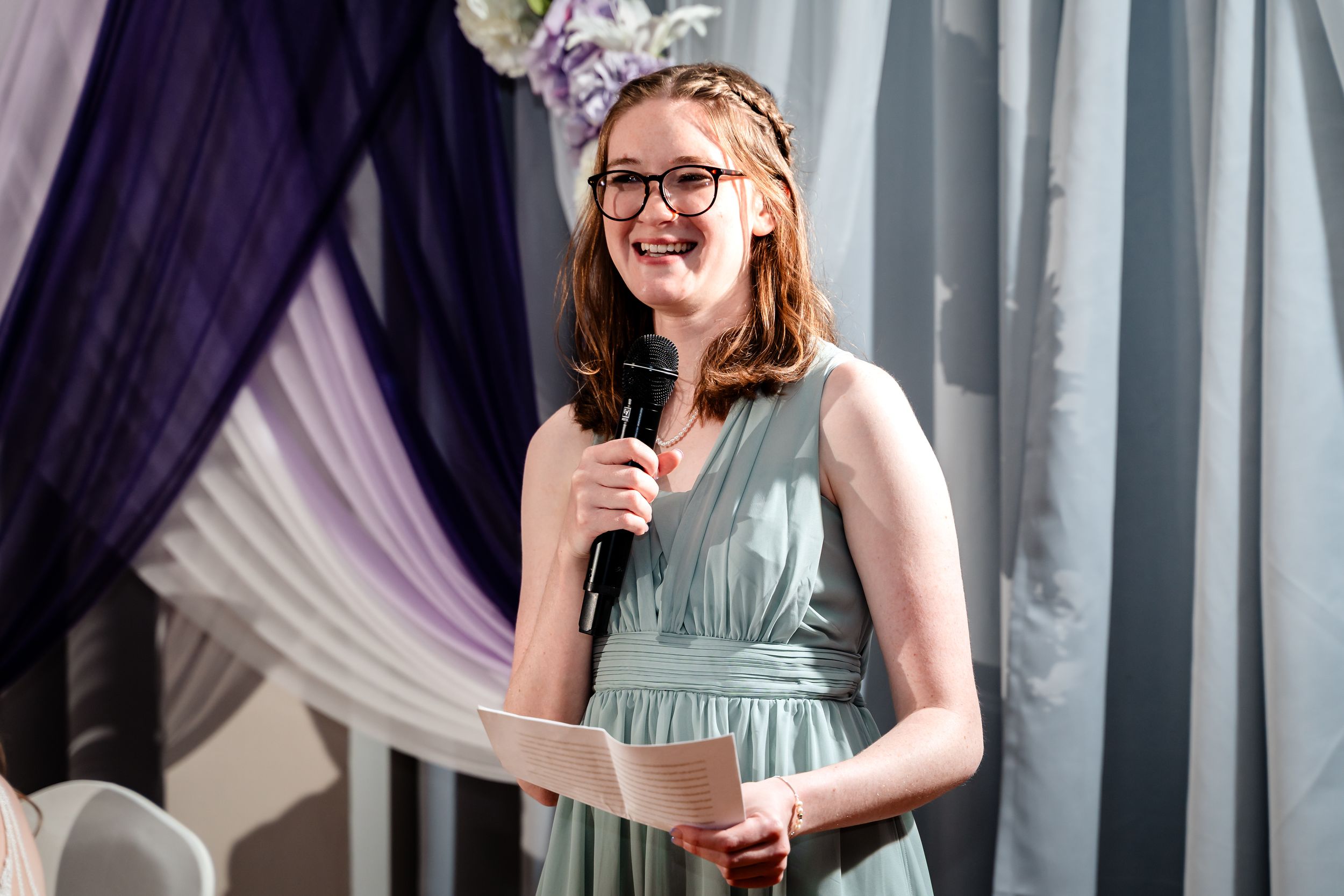 Young woman in mint bridesmaid dress giving speech at wedding ceremony, holding microphone and notes.