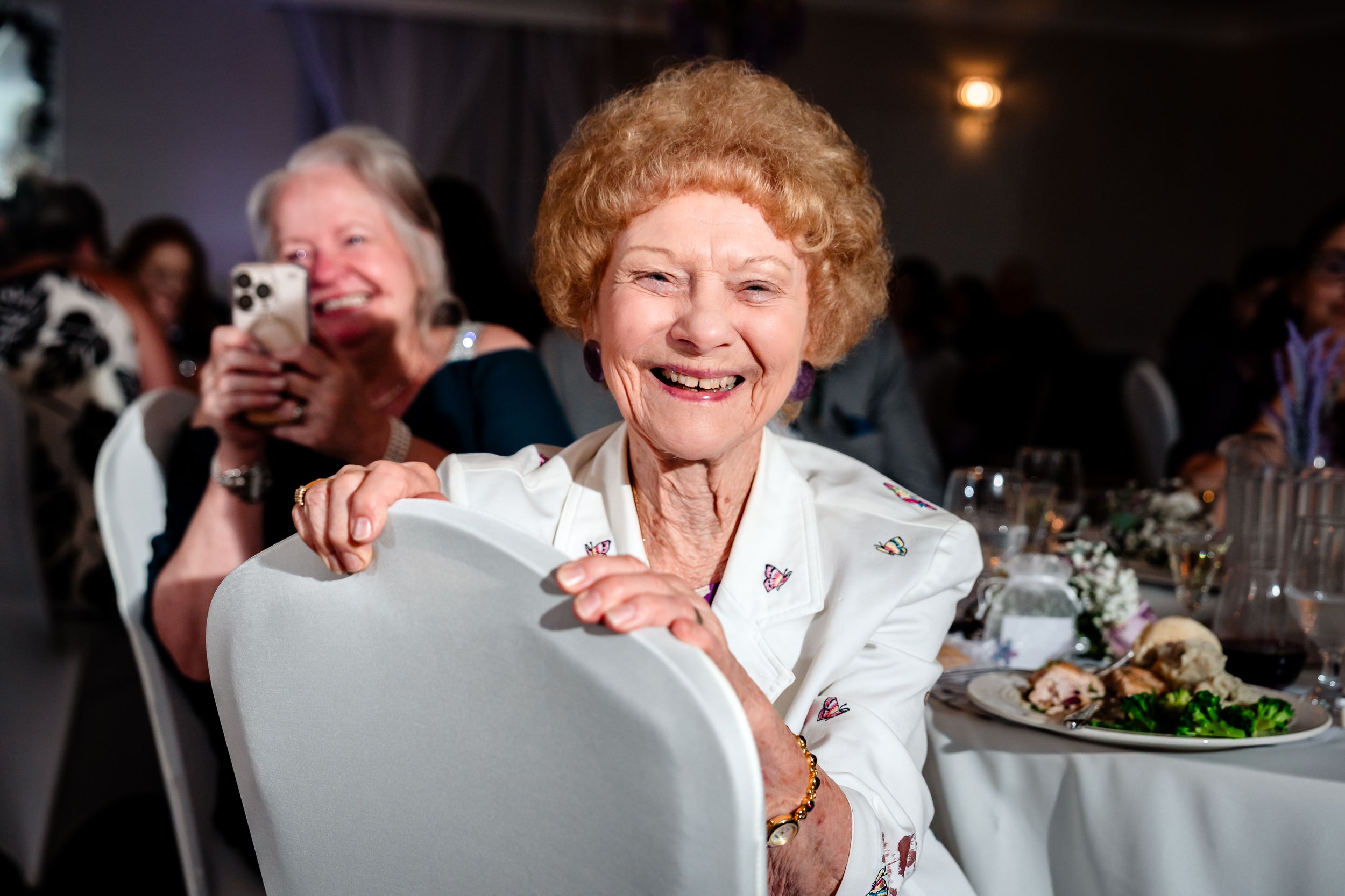 Elderly woman with red hair smiling joyfully at a formal event, seated at a decorated banquet table.