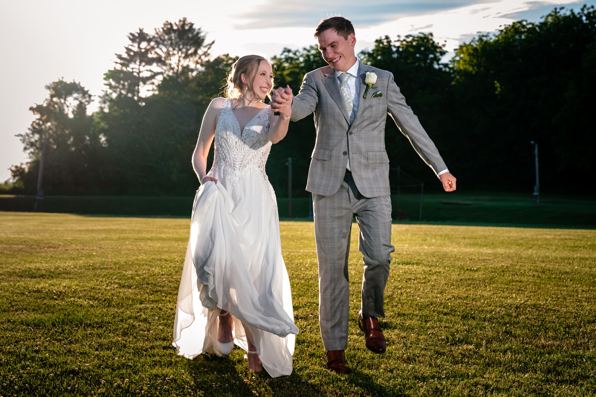 A joyful bride and groom walk hand-in-hand across a lush green lawn at golden hour during their outdoor wedding.