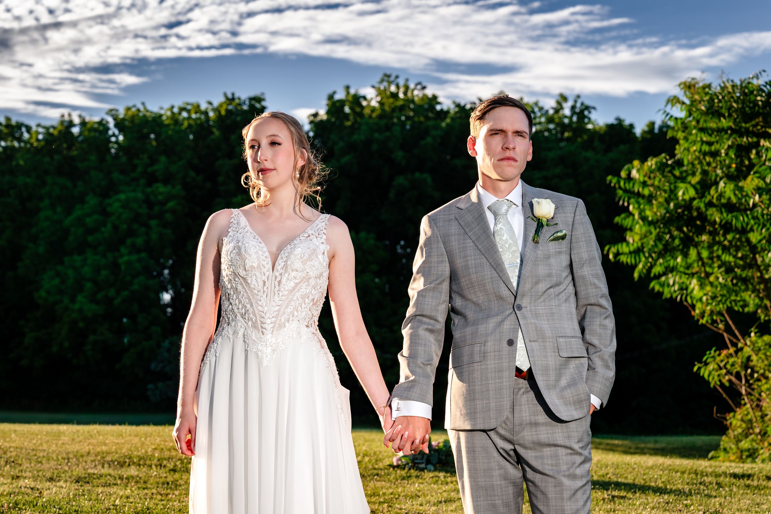 Bride in white lace gown and groom in gray suit hold hands outdoors on green lawn under blue sky.