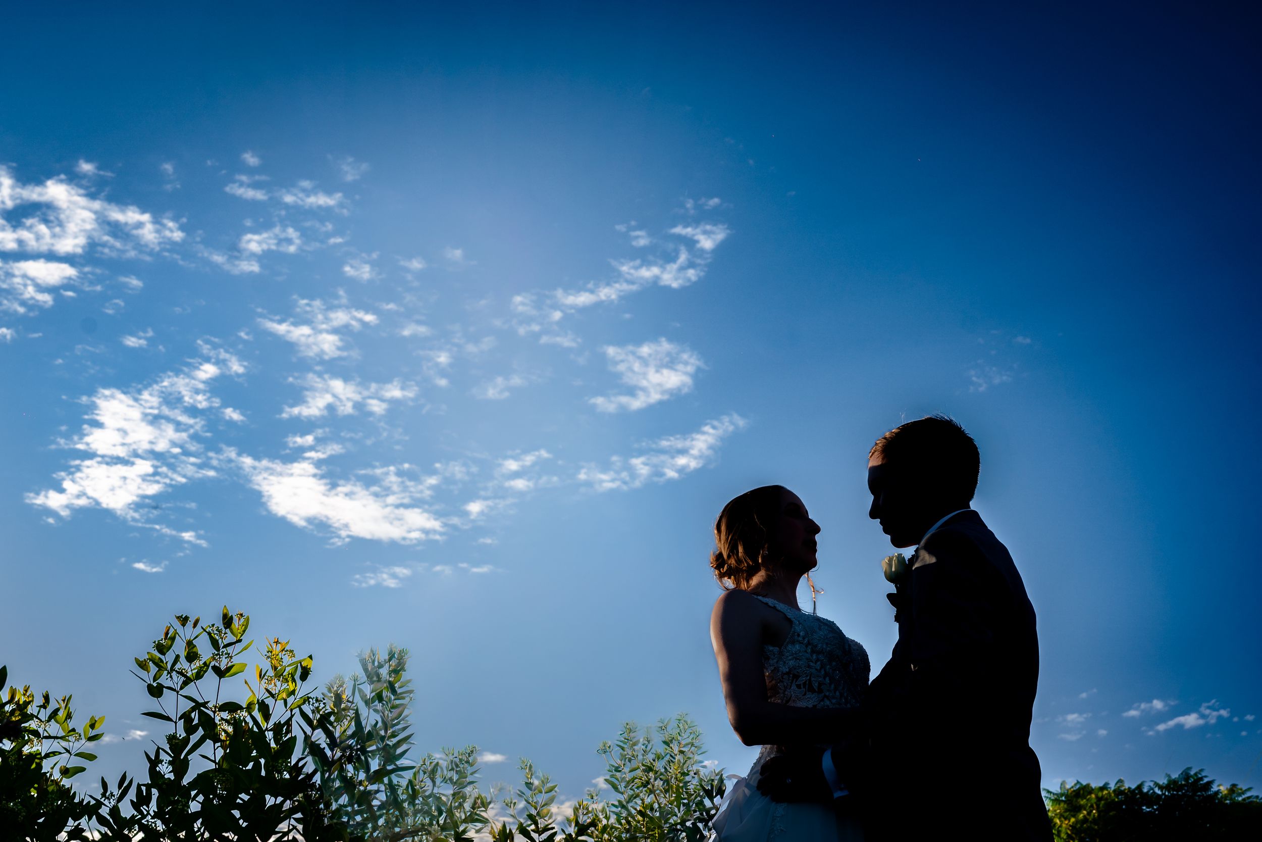 Silhouette of a bride and groom embracing against a vibrant blue sky with wispy clouds on their wedding day.