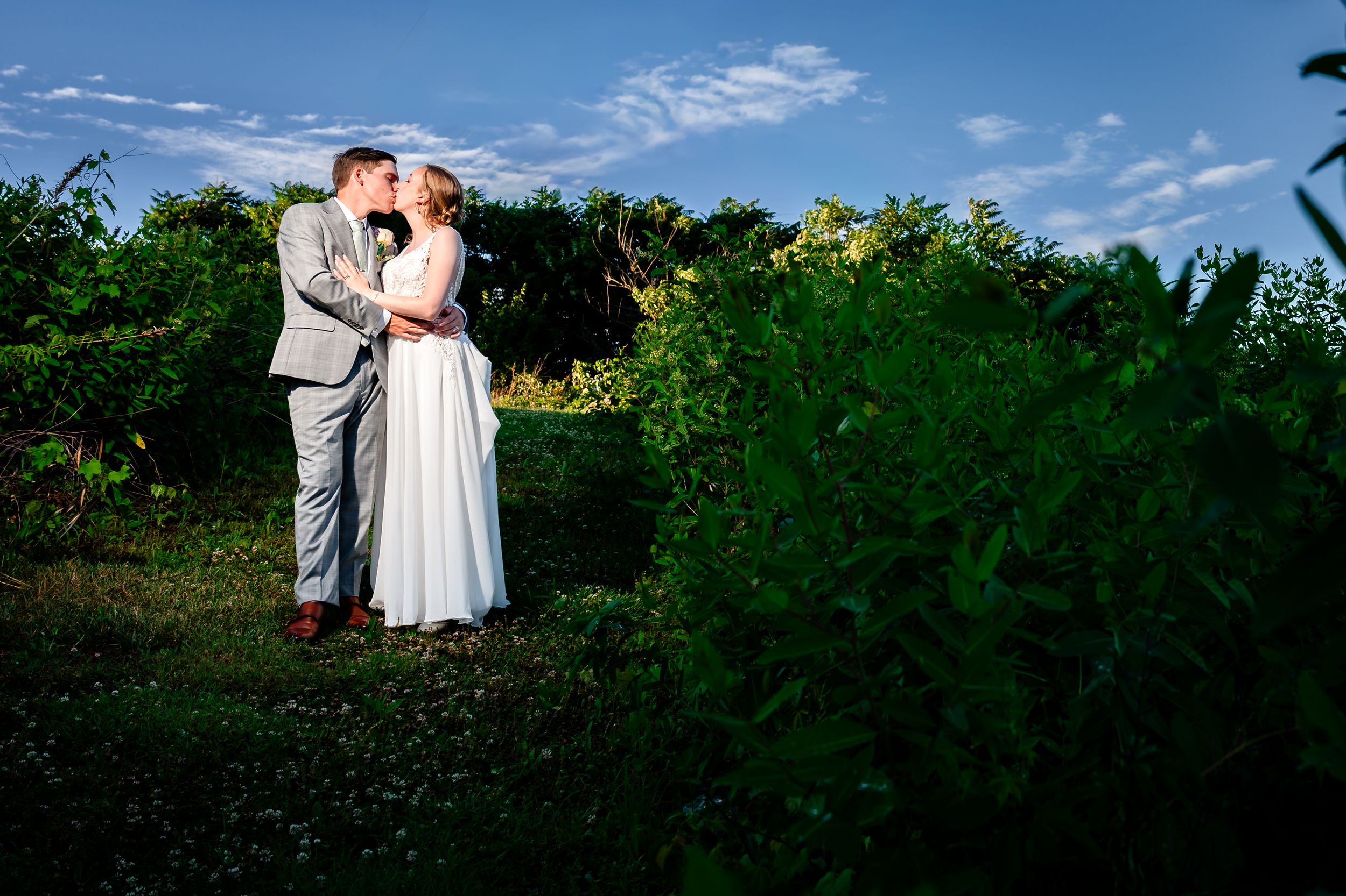 Bride and groom share a romantic kiss surrounded by lush green garden foliage on their wedding day.