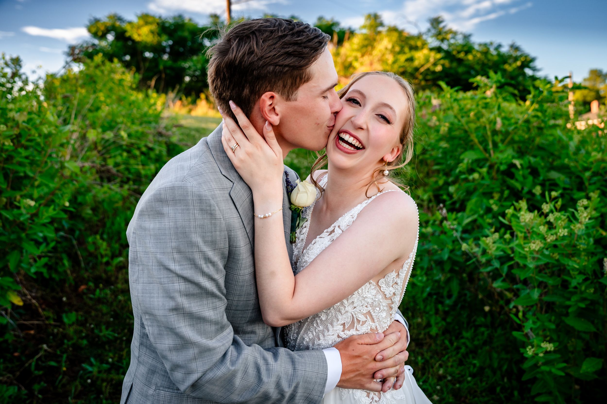 A happy bride laughs as her groom kisses her cheek outdoors at golden hour.