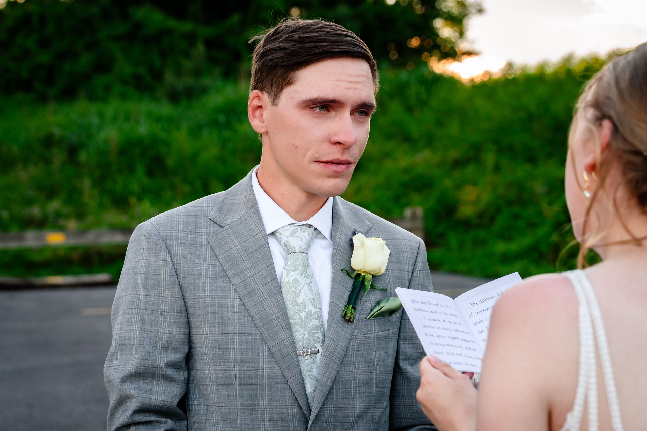 Emotional groom in gray suit listens to bride read wedding vows outdoors during intimate ceremony.