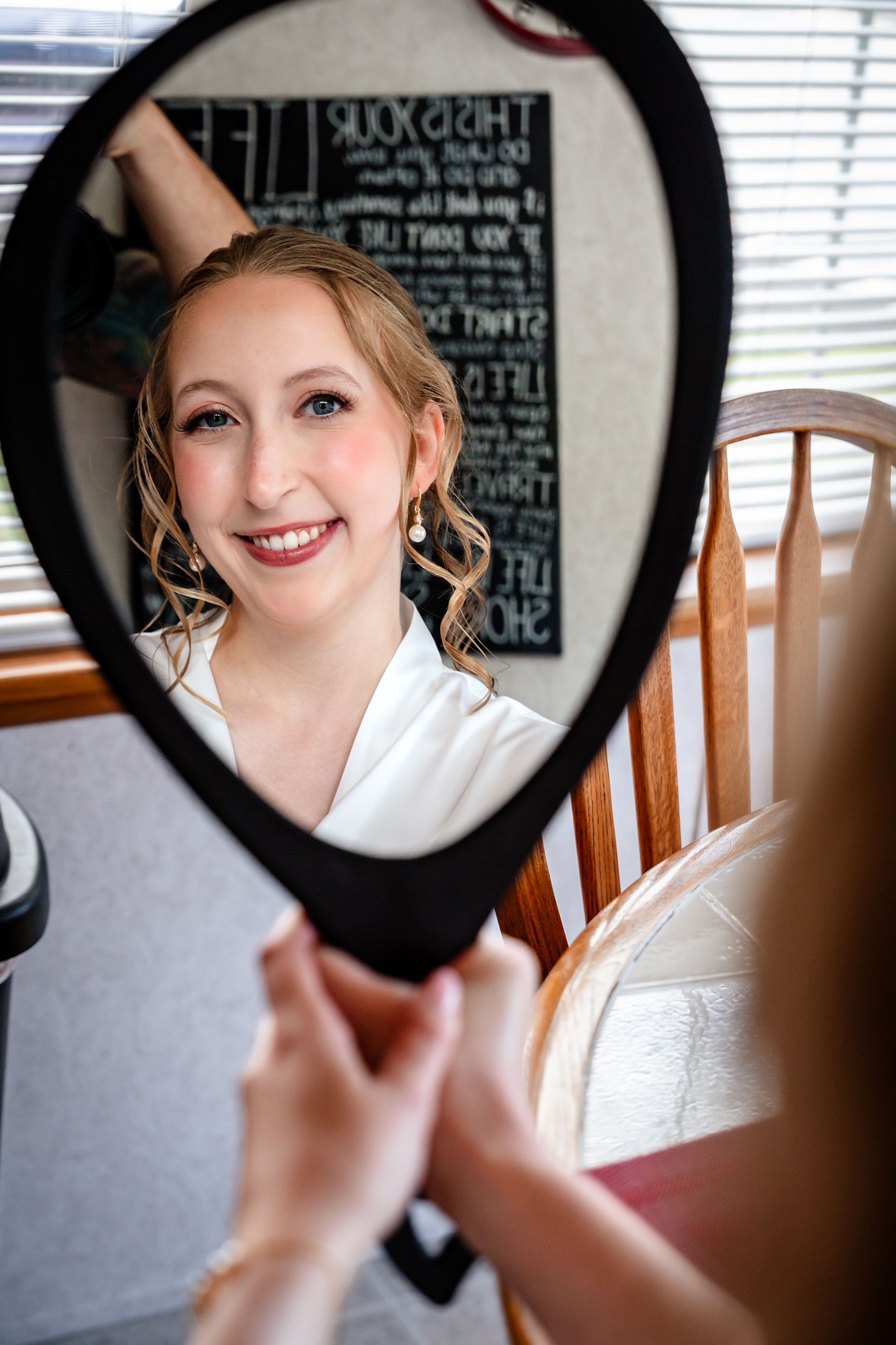 Radiant bride admires her makeup in handheld mirror during wedding day preparations.