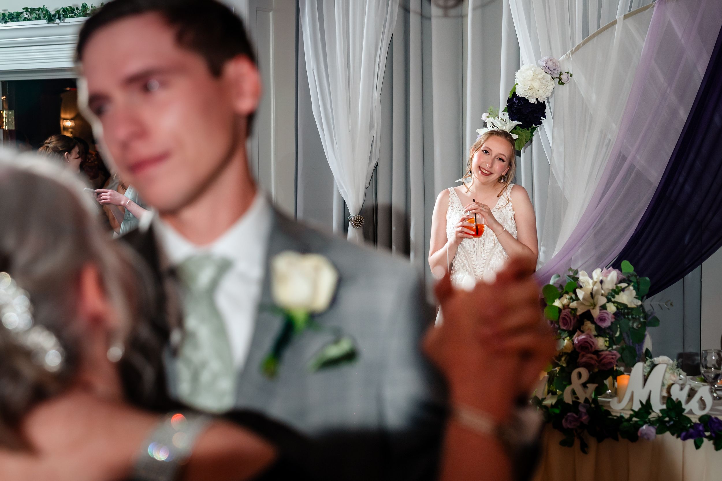 Bride smiling with a drink watches groom dance with guest at elegant wedding reception with floral decorations.