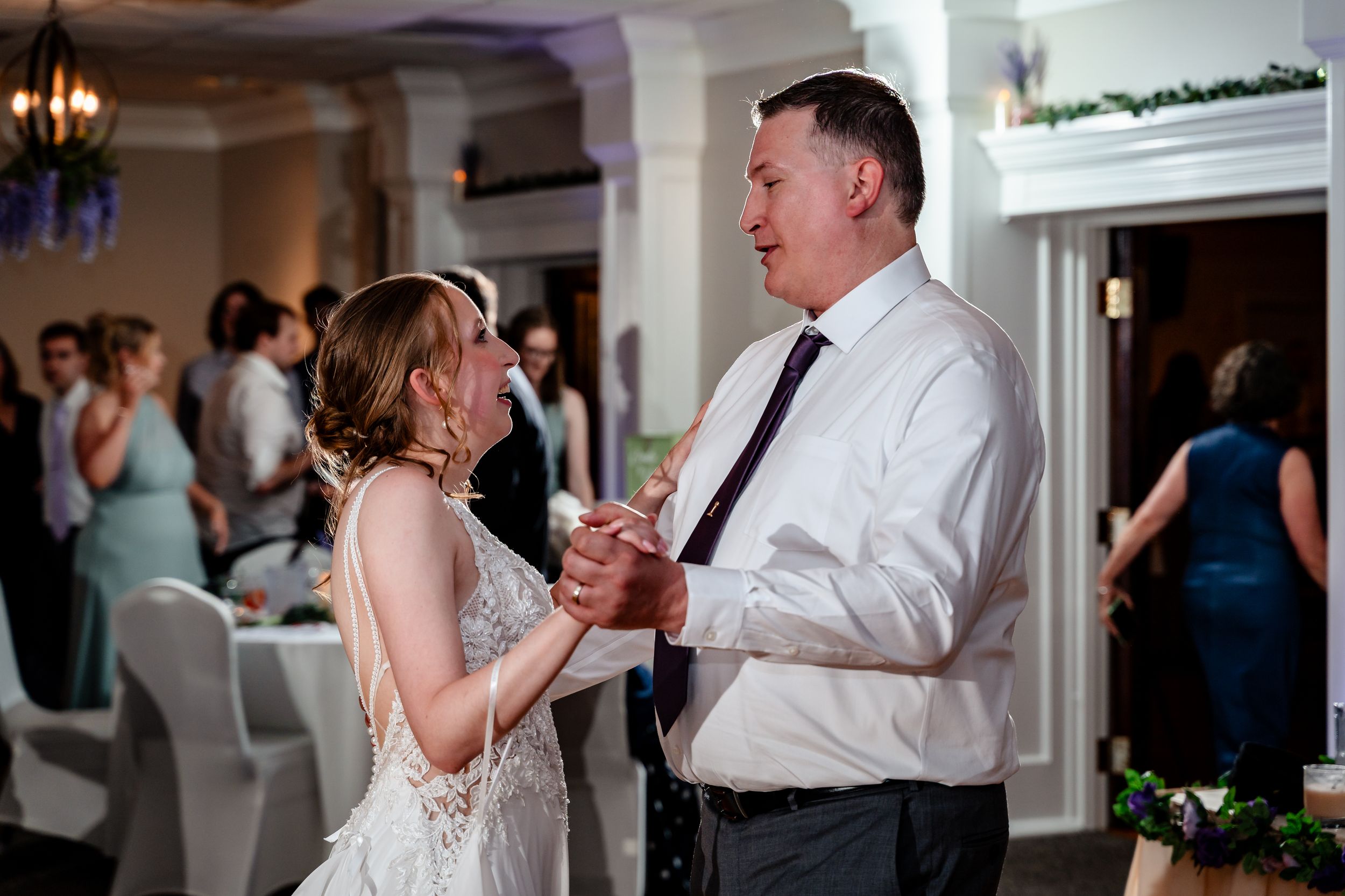 Bride and father share an emotional first dance at elegant wedding reception venue with guests in background.