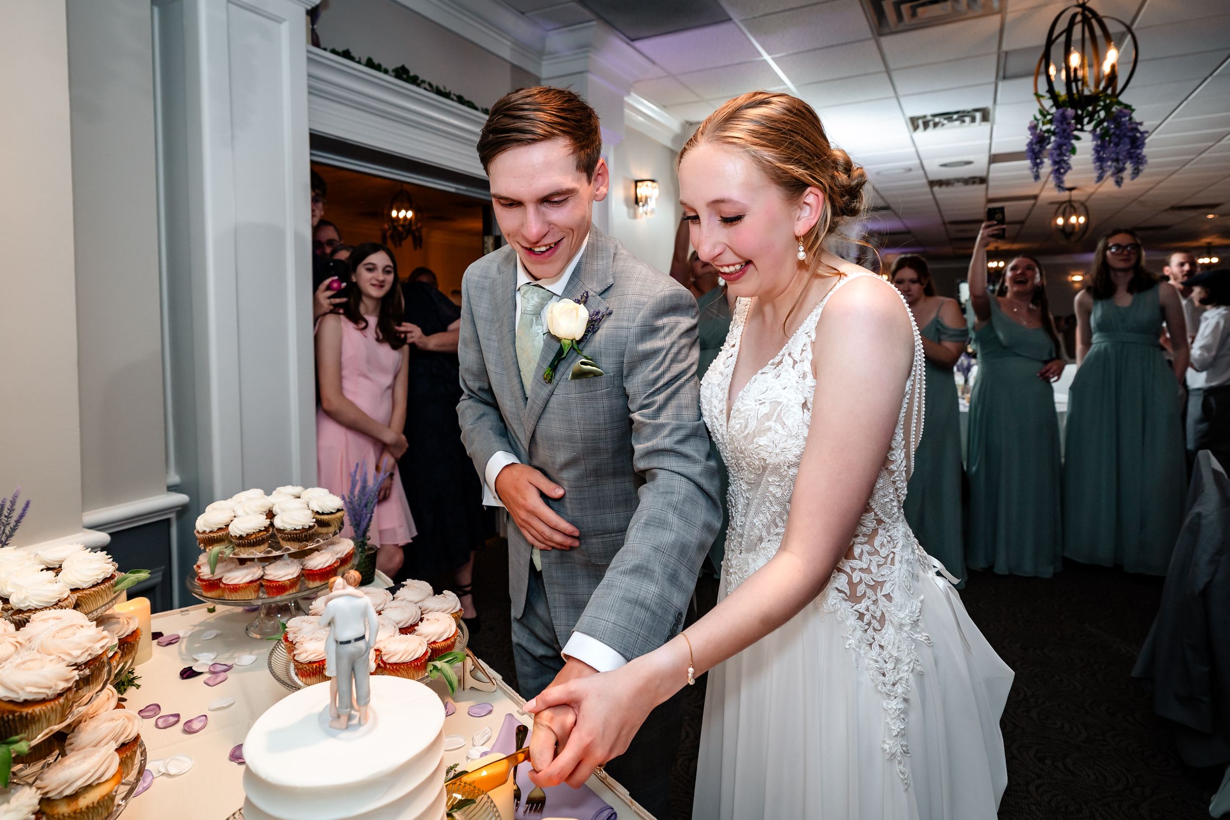 Bride and groom smiling while cutting their wedding cake at a reception with guests in the background.