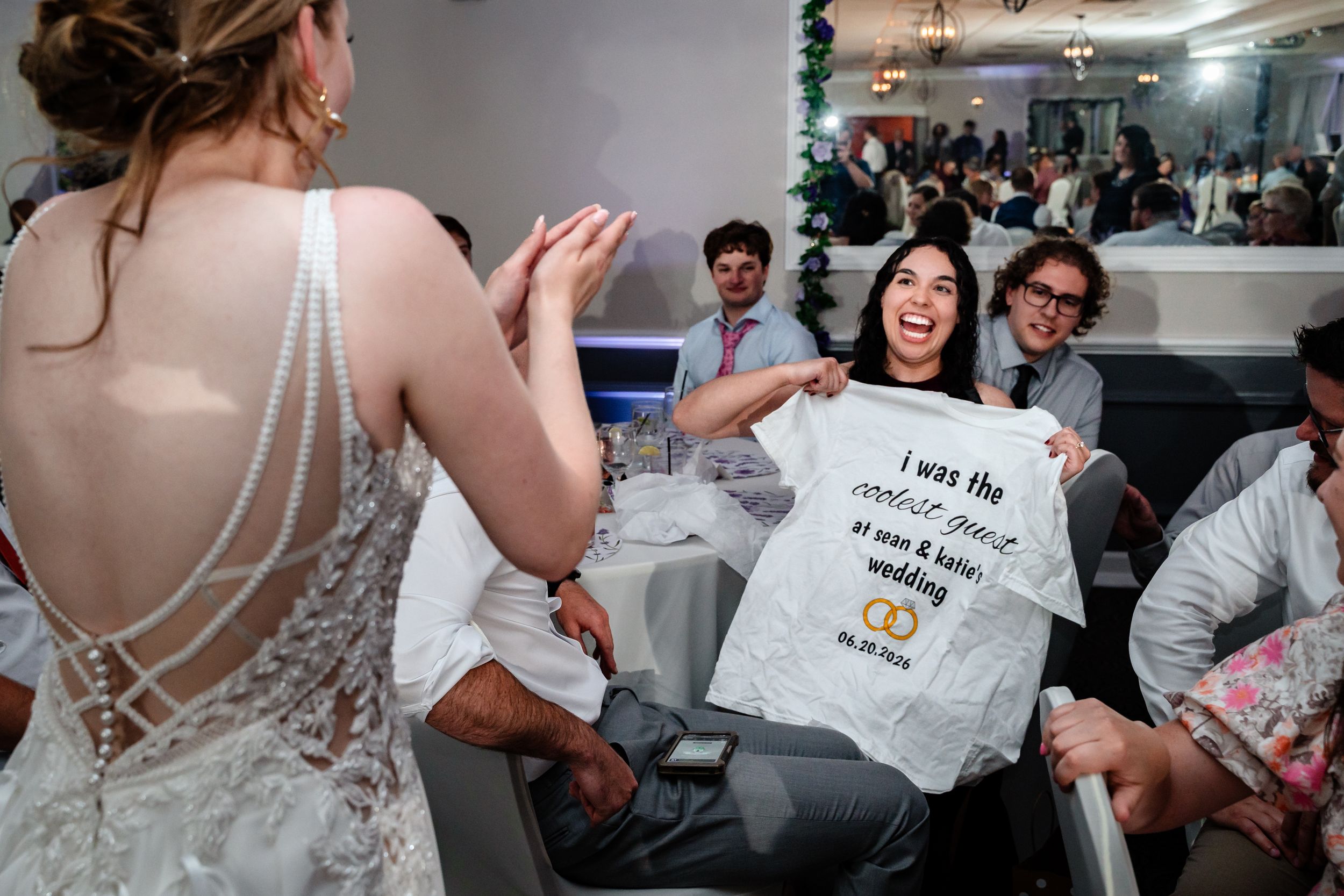 Bride showing a humorous wedding guest t-shirt at a lively indoor wedding reception celebration.