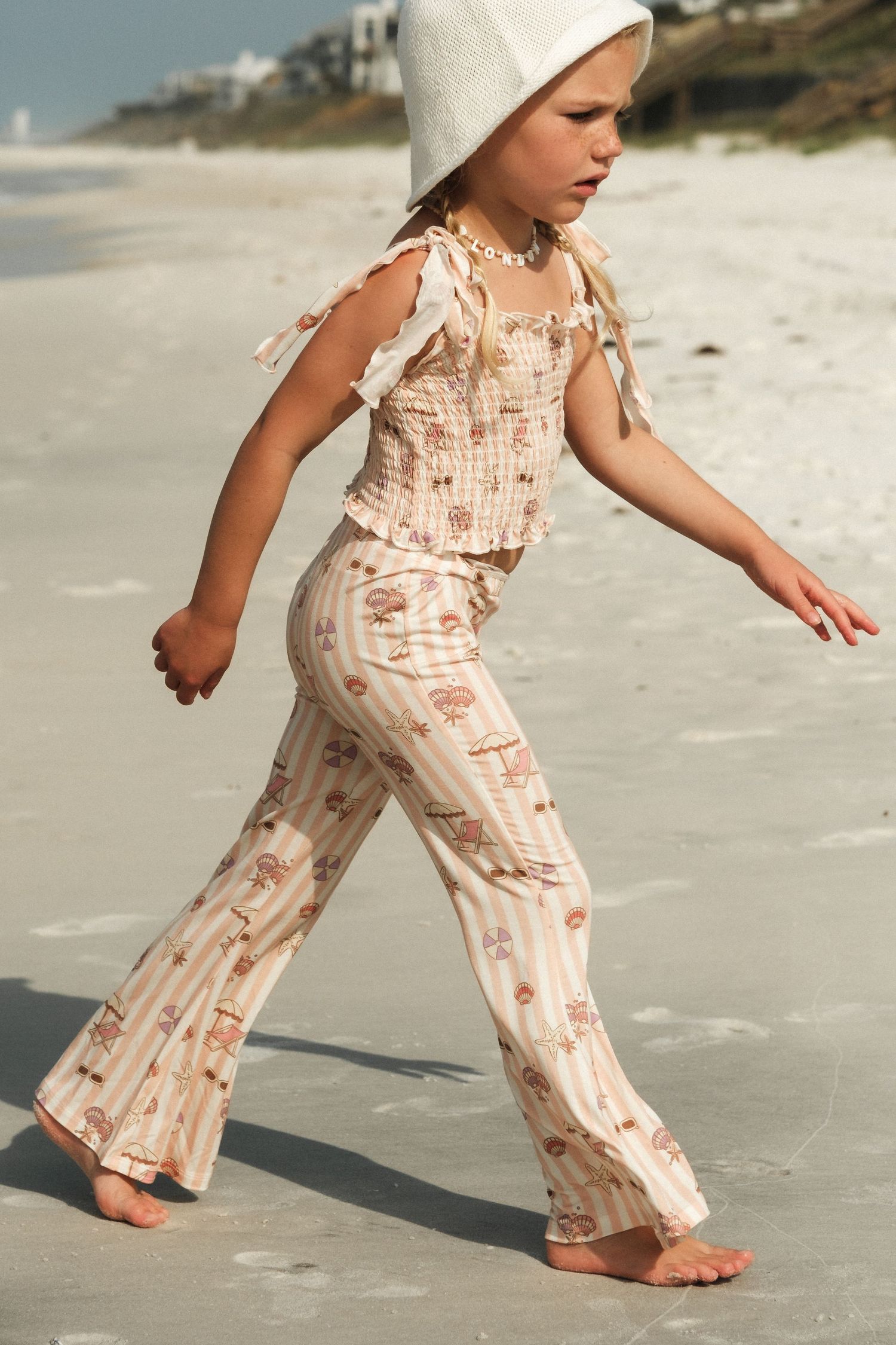 Young girl walking on beach wearing floral flare pants, smocked top, and white sun hat.