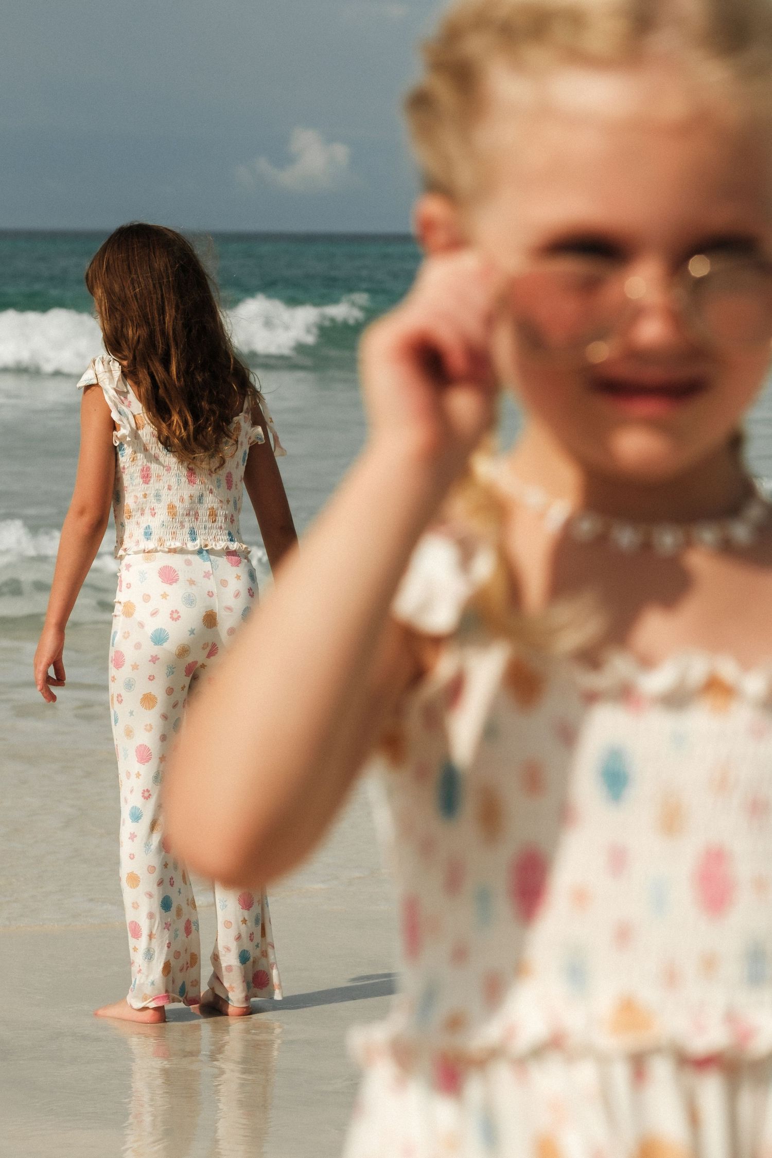 Two young girls in polka dot outfits enjoy a sunny beach day, one walking toward ocean waves while the other poses in the foreground.