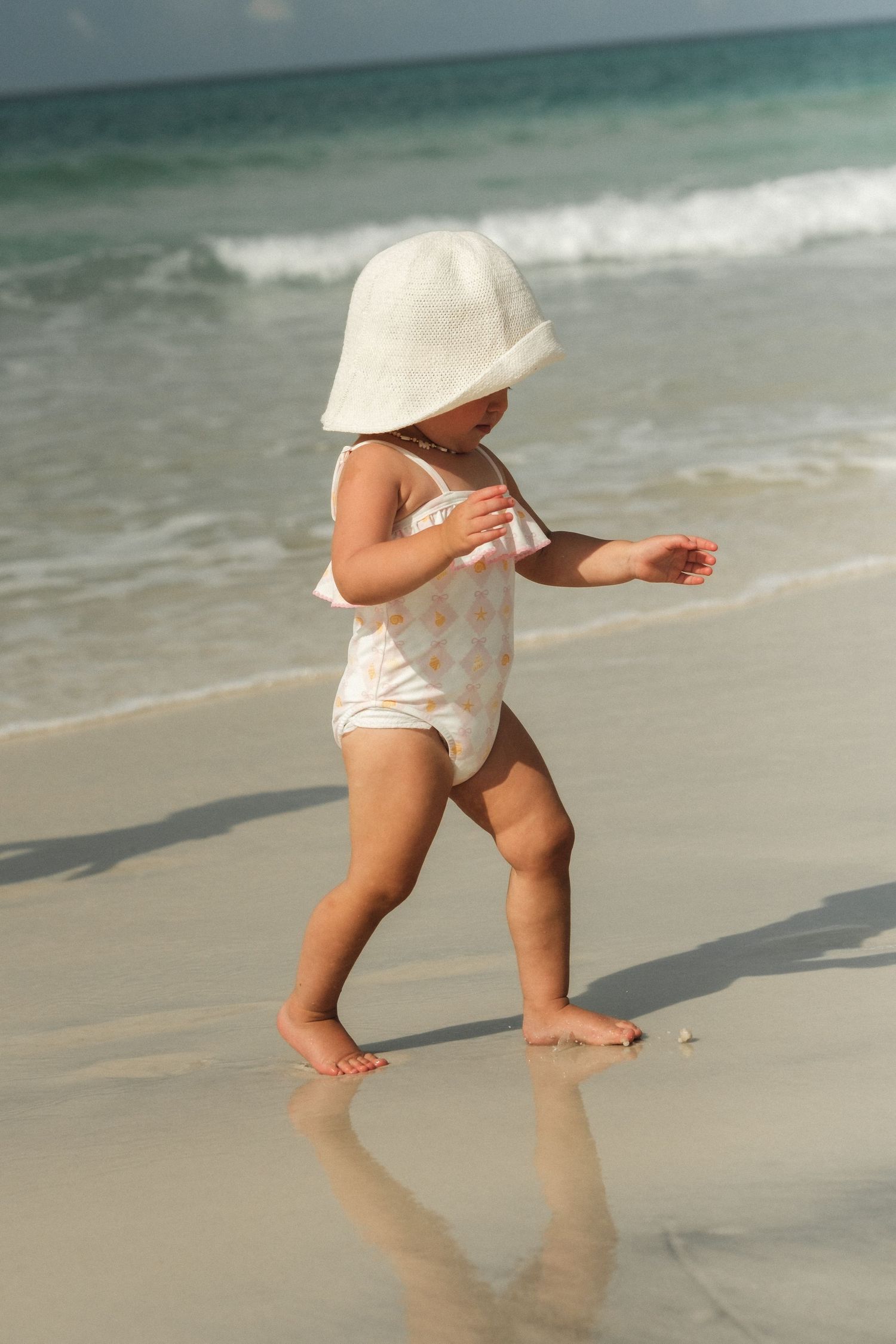 A toddler in a white sun hat and swimsuit walks barefoot along a sandy beach with ocean waves in the background.