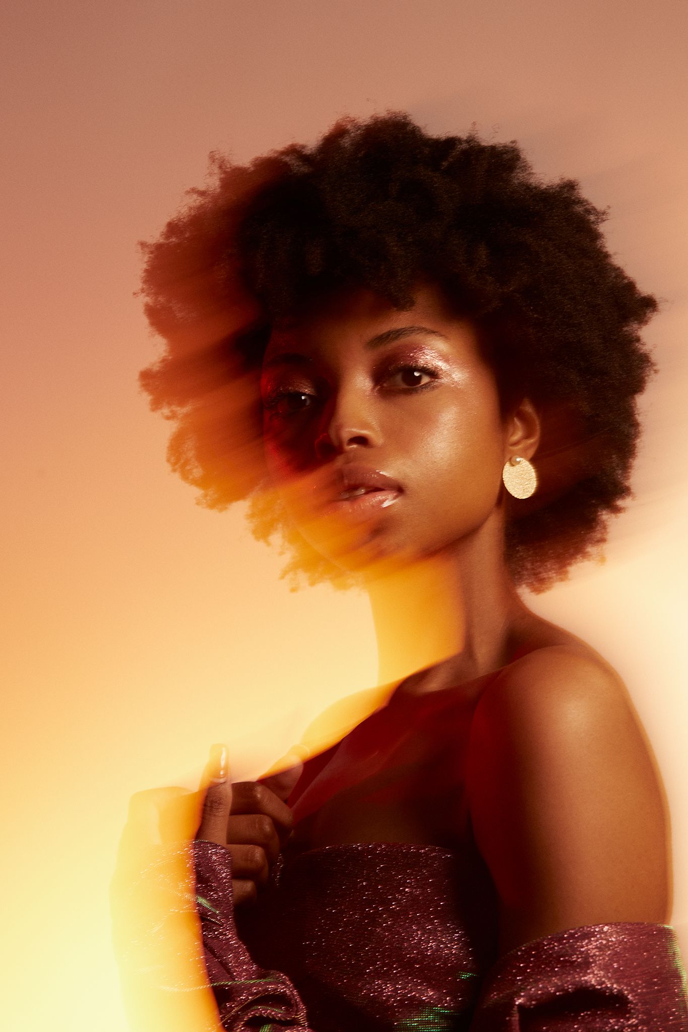 A stunning Black woman with a voluminous afro poses in a purple sequin dress under warm golden studio lighting.