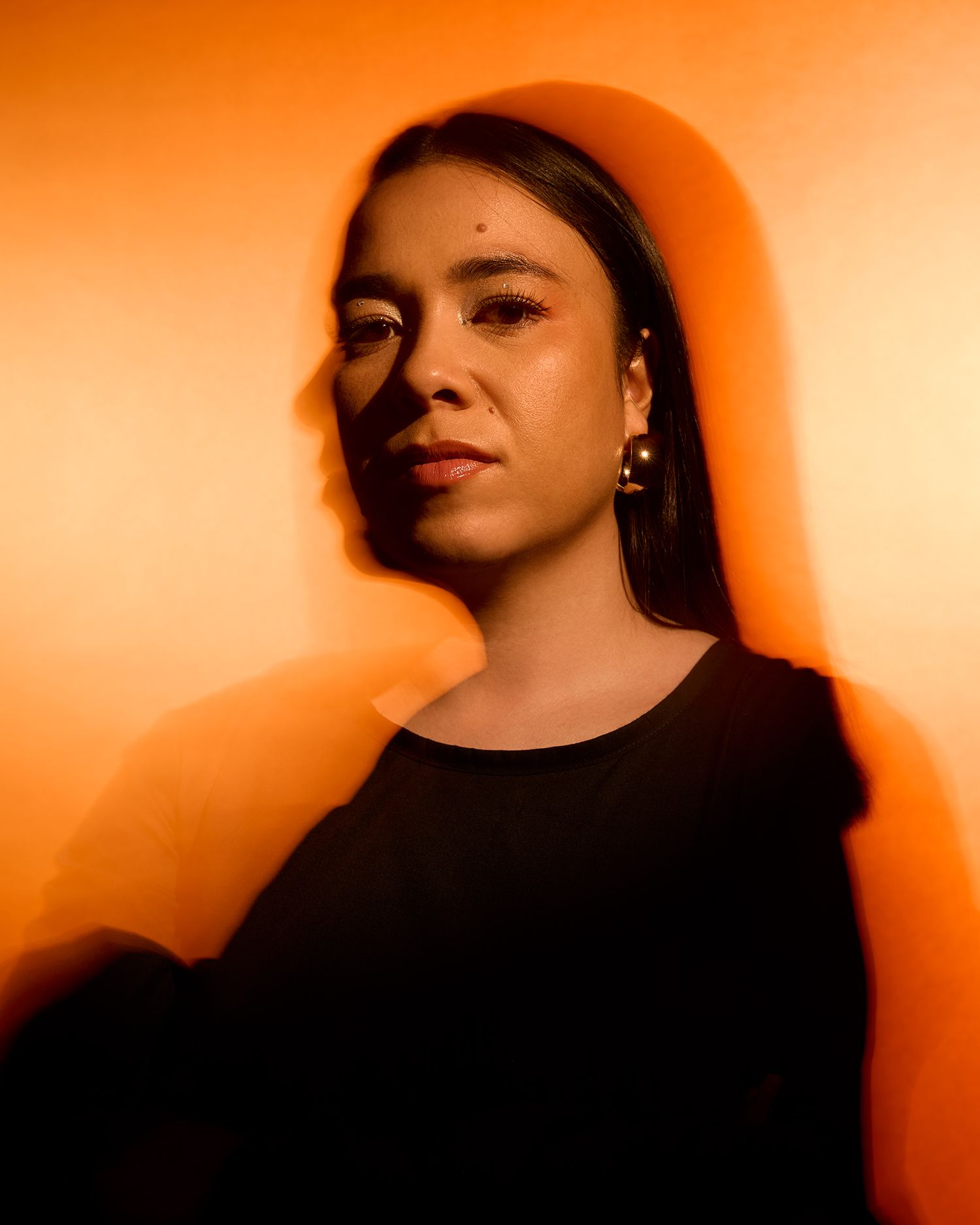 Portrait of a woman with dark hair and gold earrings bathed in warm orange light.