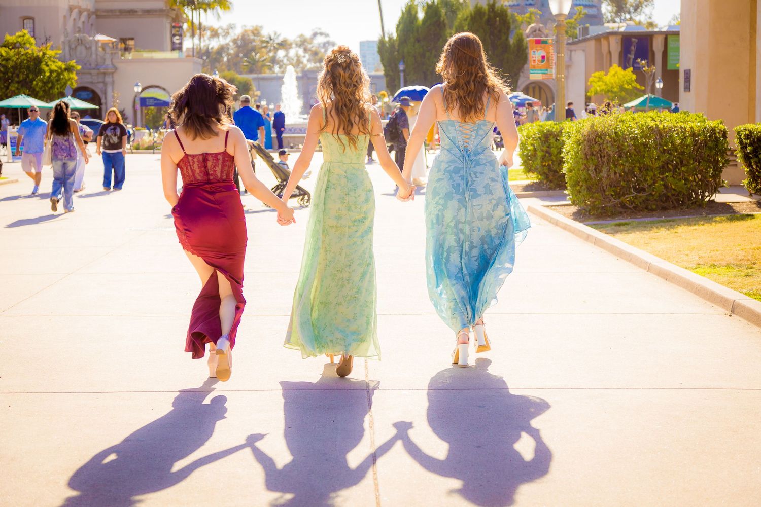 Three women in colorful dresses walk hand-in-hand through a sunny theme park pathway.