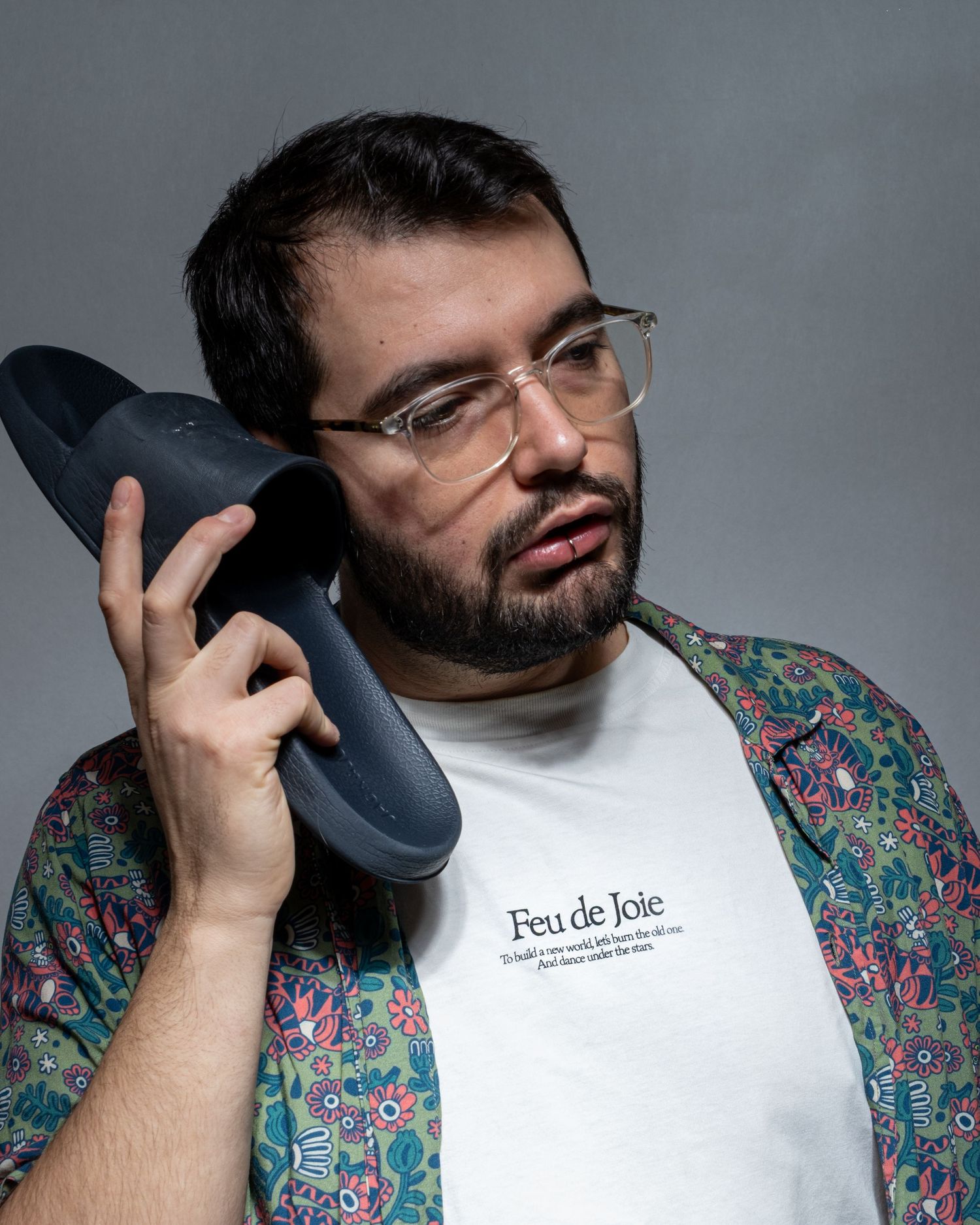Portrait masculin en studio tenant une chaussure comme un téléphone dans une mise en scène décalée.