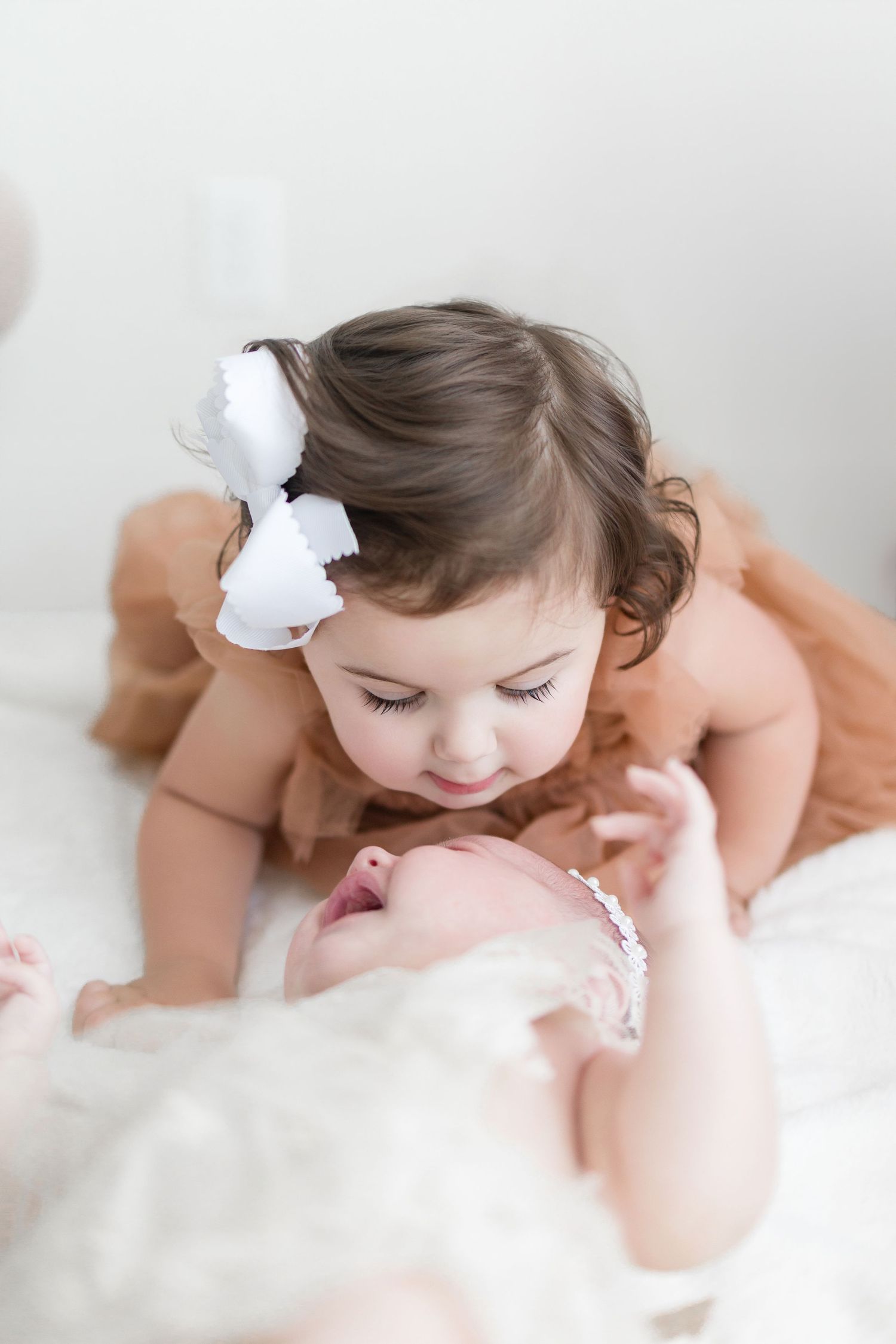 Toddler girl in peach dress with white bow gazes lovingly at newborn baby sibling during professional photo session.