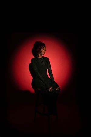 Woman in black outfit seated on stool against dramatic red spotlight background in dark studio setting.